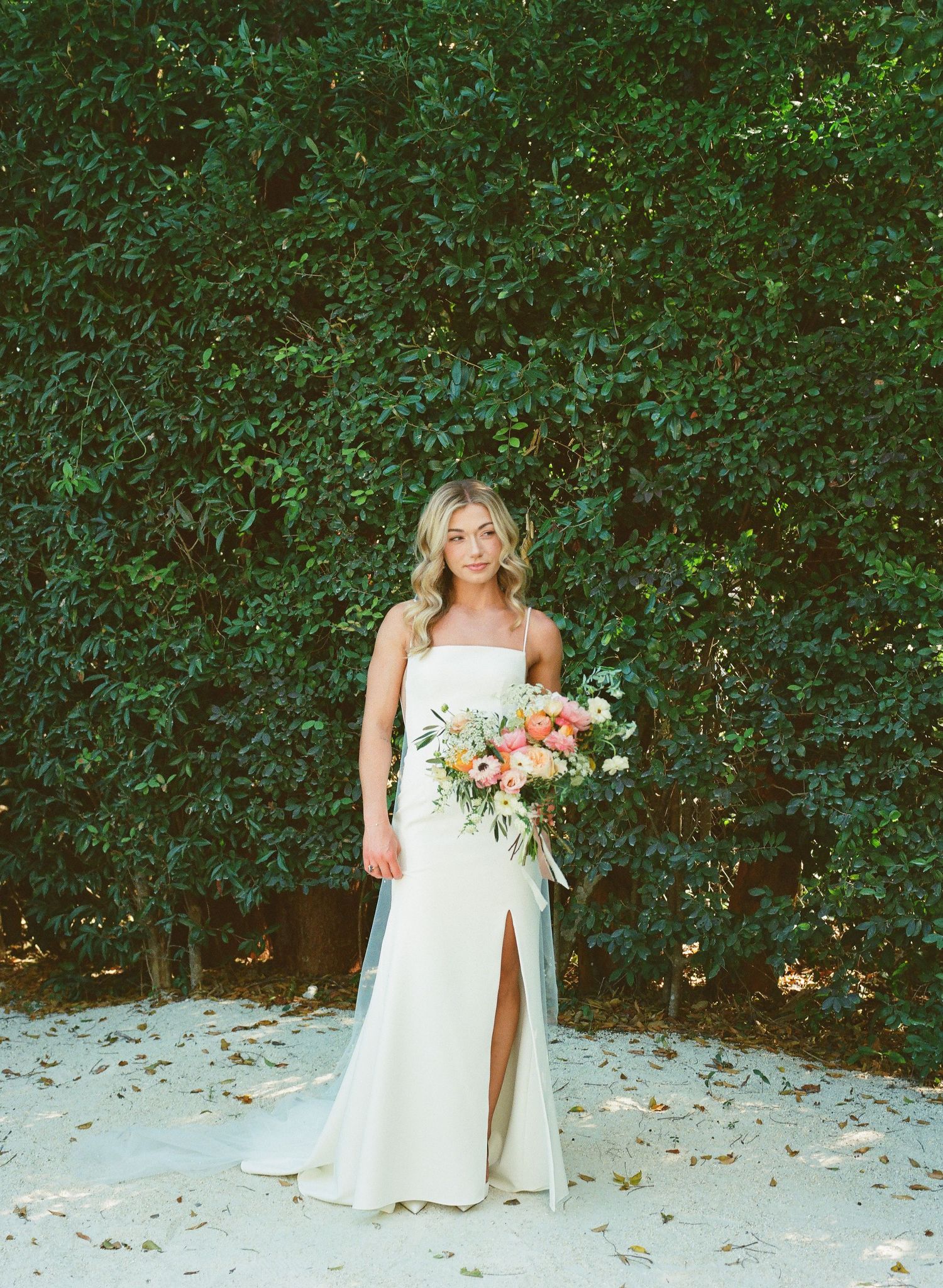 Bridal portrait with peach and coral bouquet against lush green hedge background.