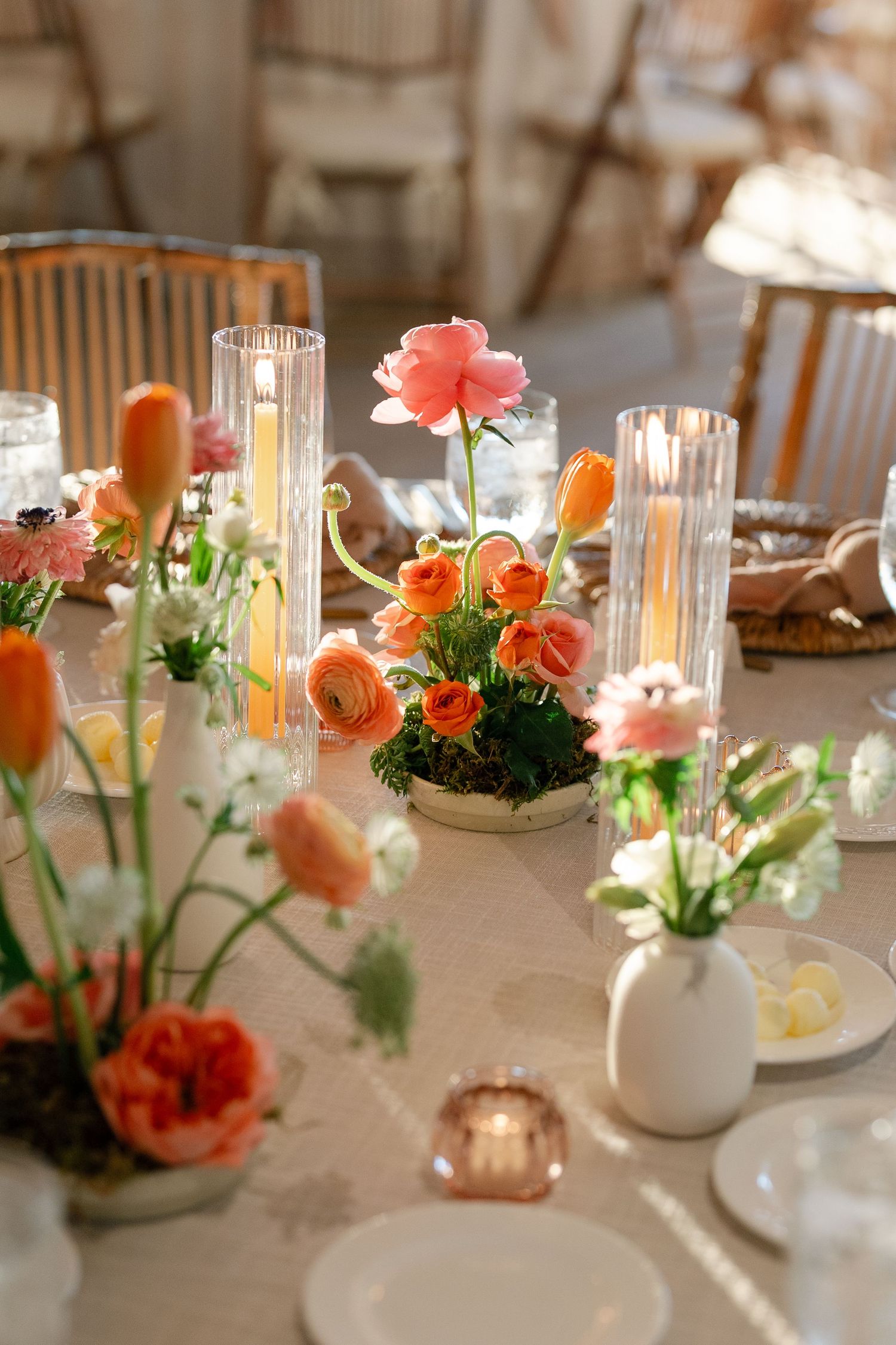 Elegant table setting with coral and peach flowers in white vases alongside glowing glass candleholders on a light tablecloth.