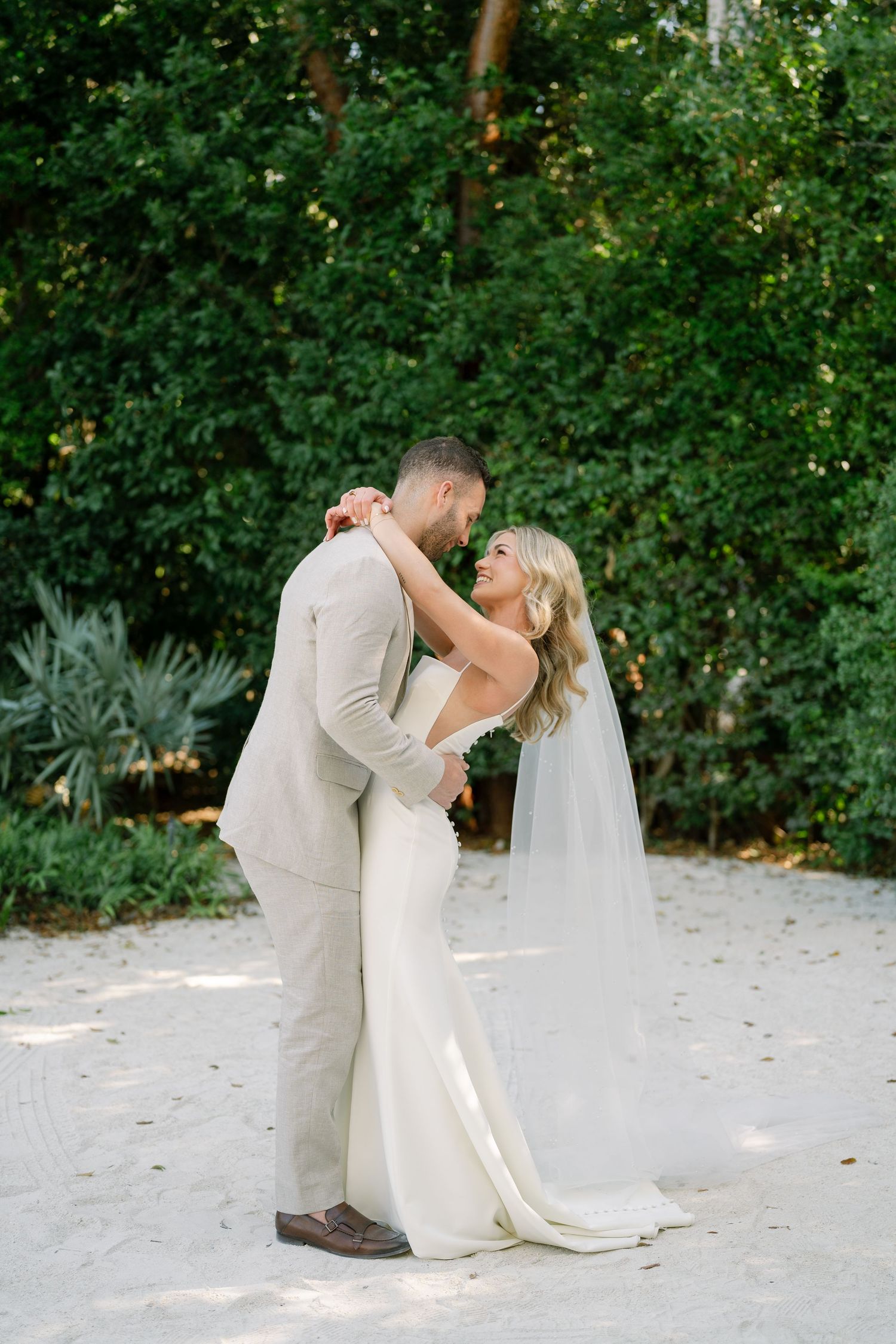 Romantic first dance moment between newlyweds on sandy beach surrounded by greenery.