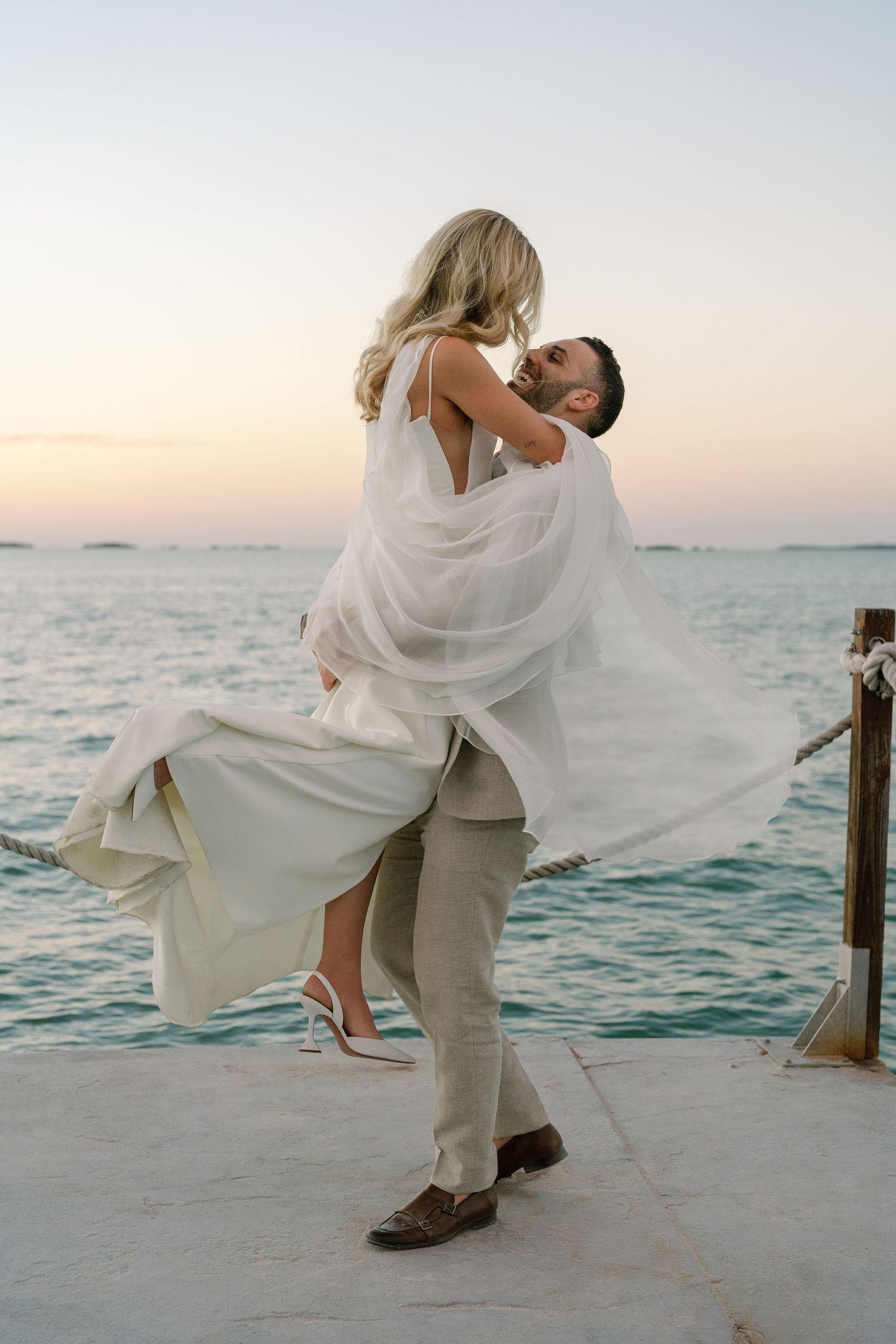A romantic dip pose between newlyweds on a waterfront dock at sunset.