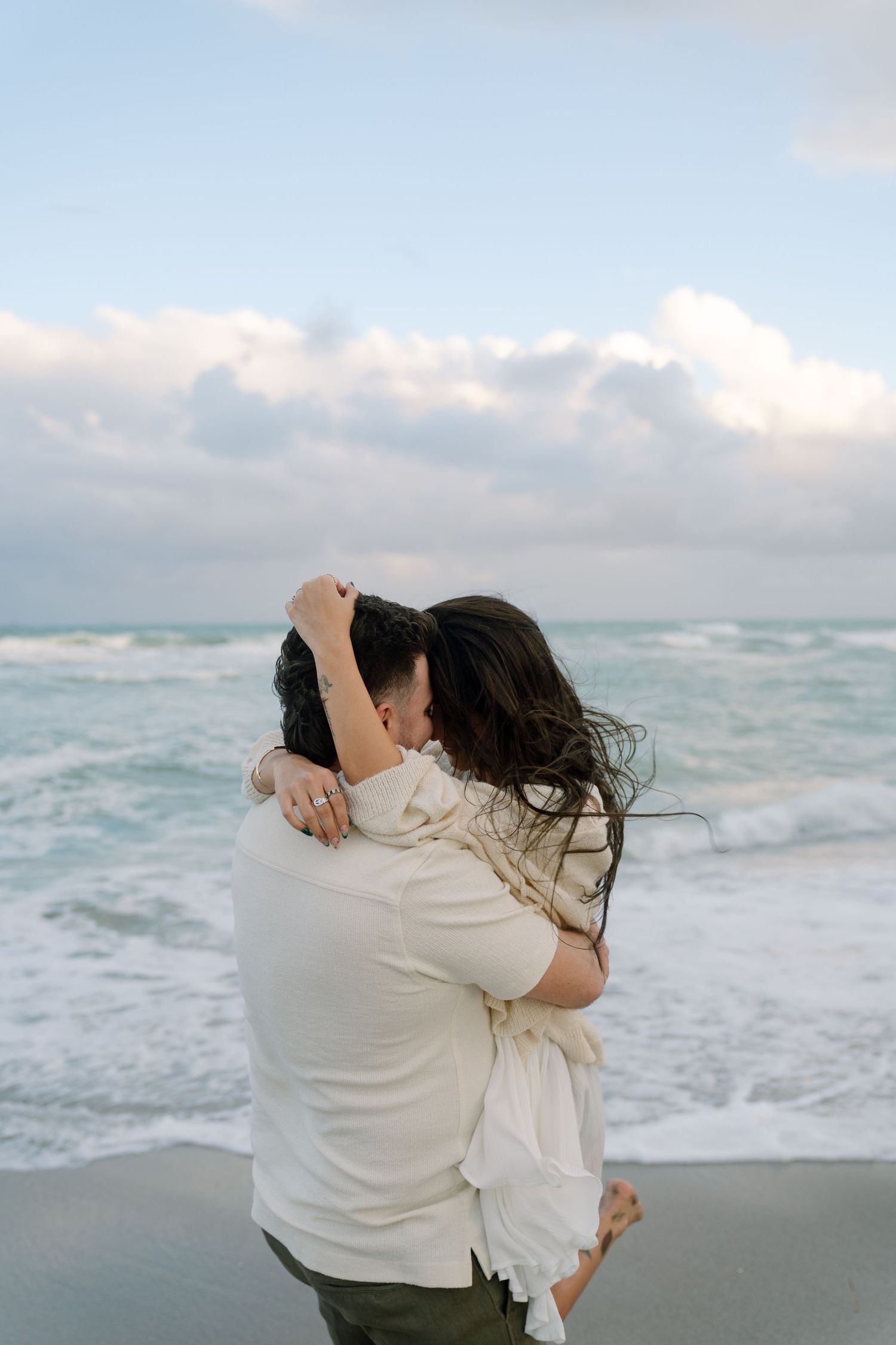A couple embraces passionately on a sandy beach with waves and cloudy skies in the background.