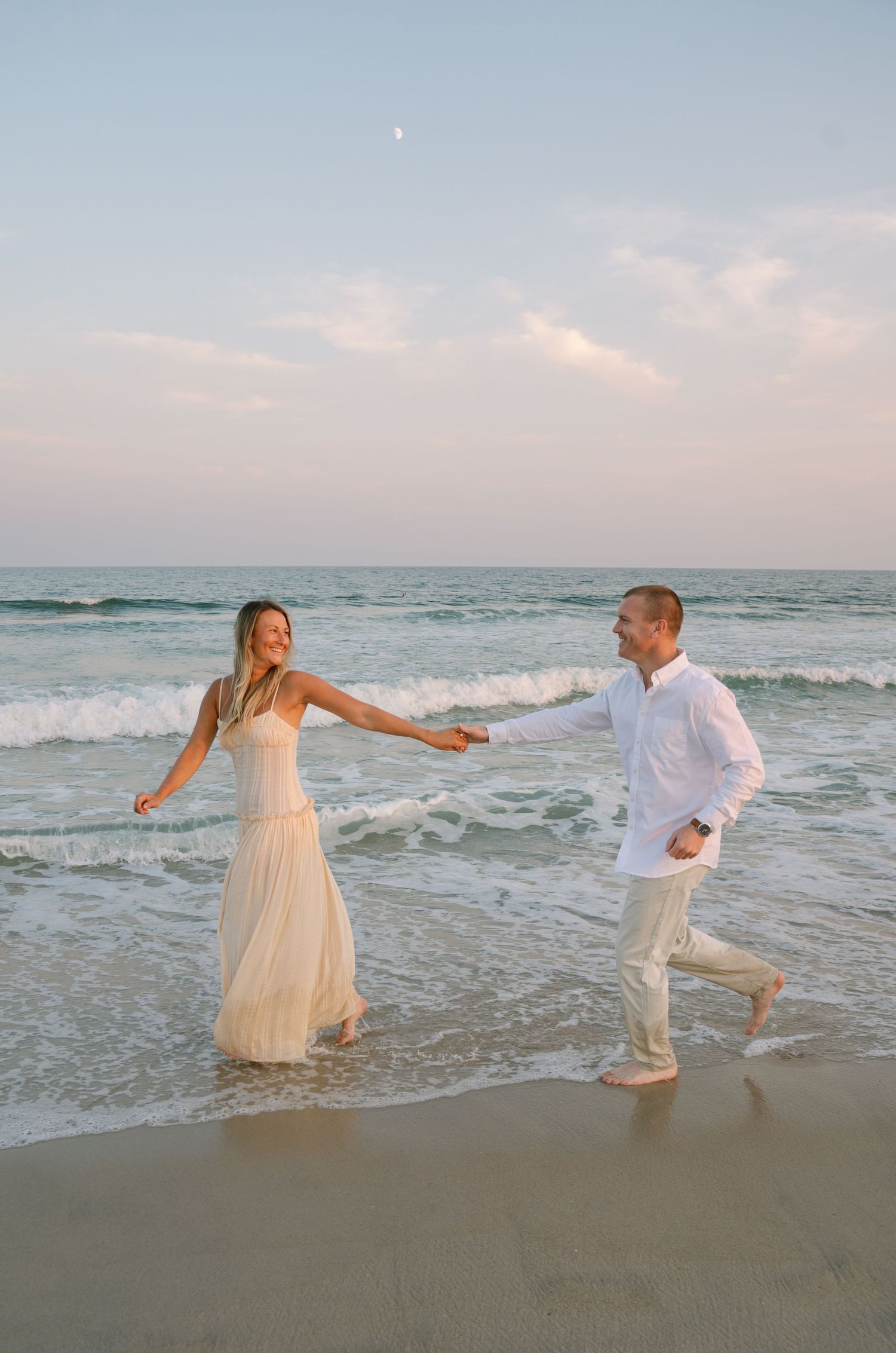 A couple in white outfits runs joyfully along the beach at sunset with ocean waves in the background.