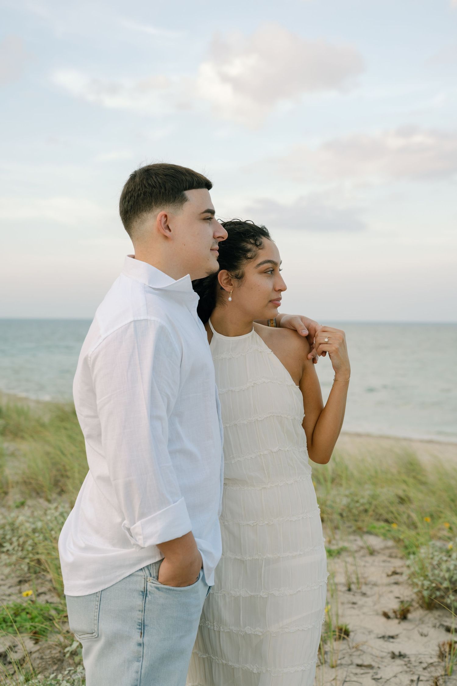 A romantic couple shares a tender moment on a beach with ocean waves and grass in the background.