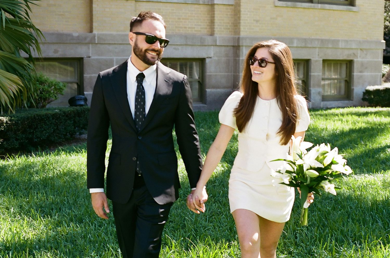A couple in formal wedding attire walks together holding hands on a sunny lawn with trees in the background.