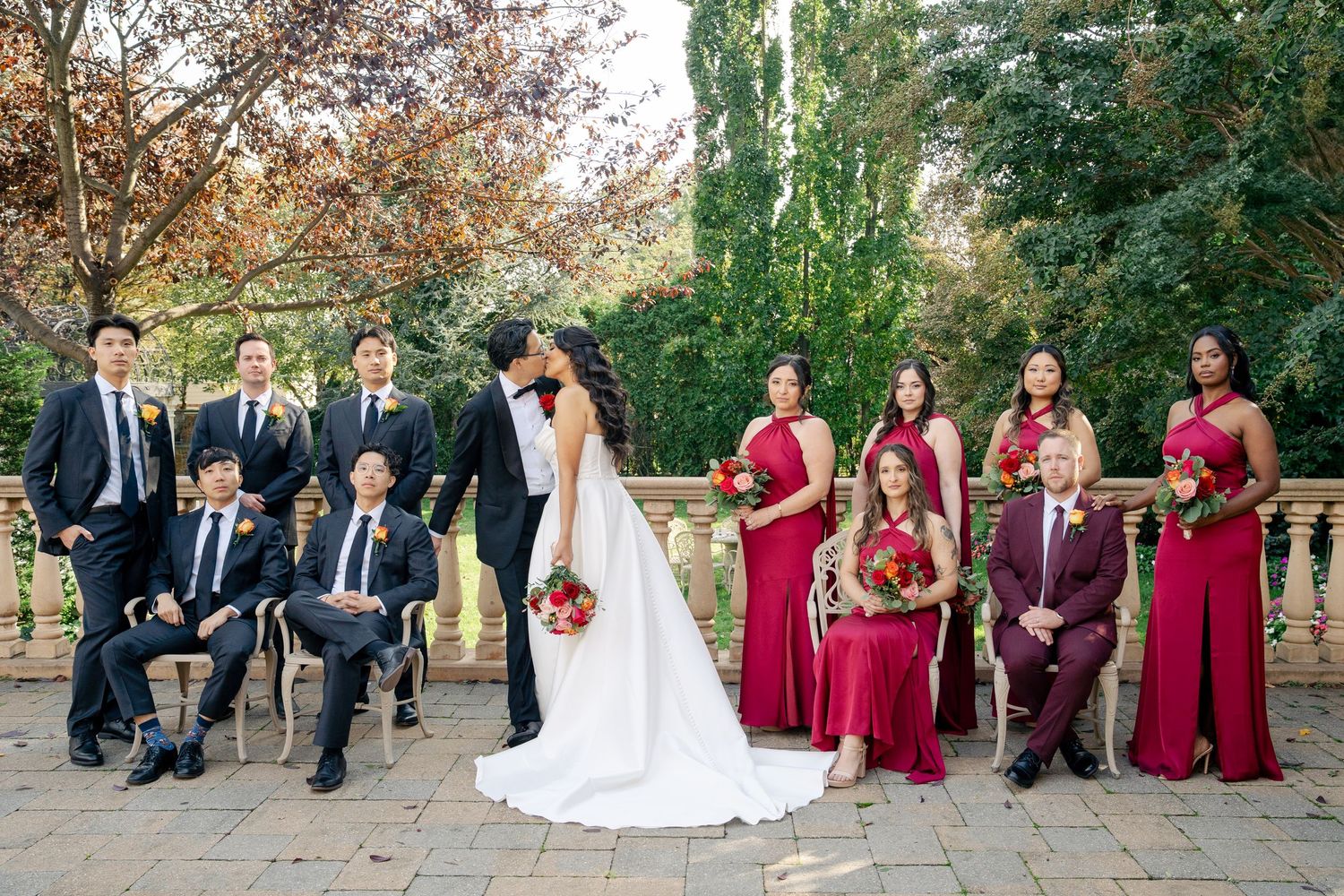 Wedding party in burgundy and navy attire poses together in an outdoor garden setting.
