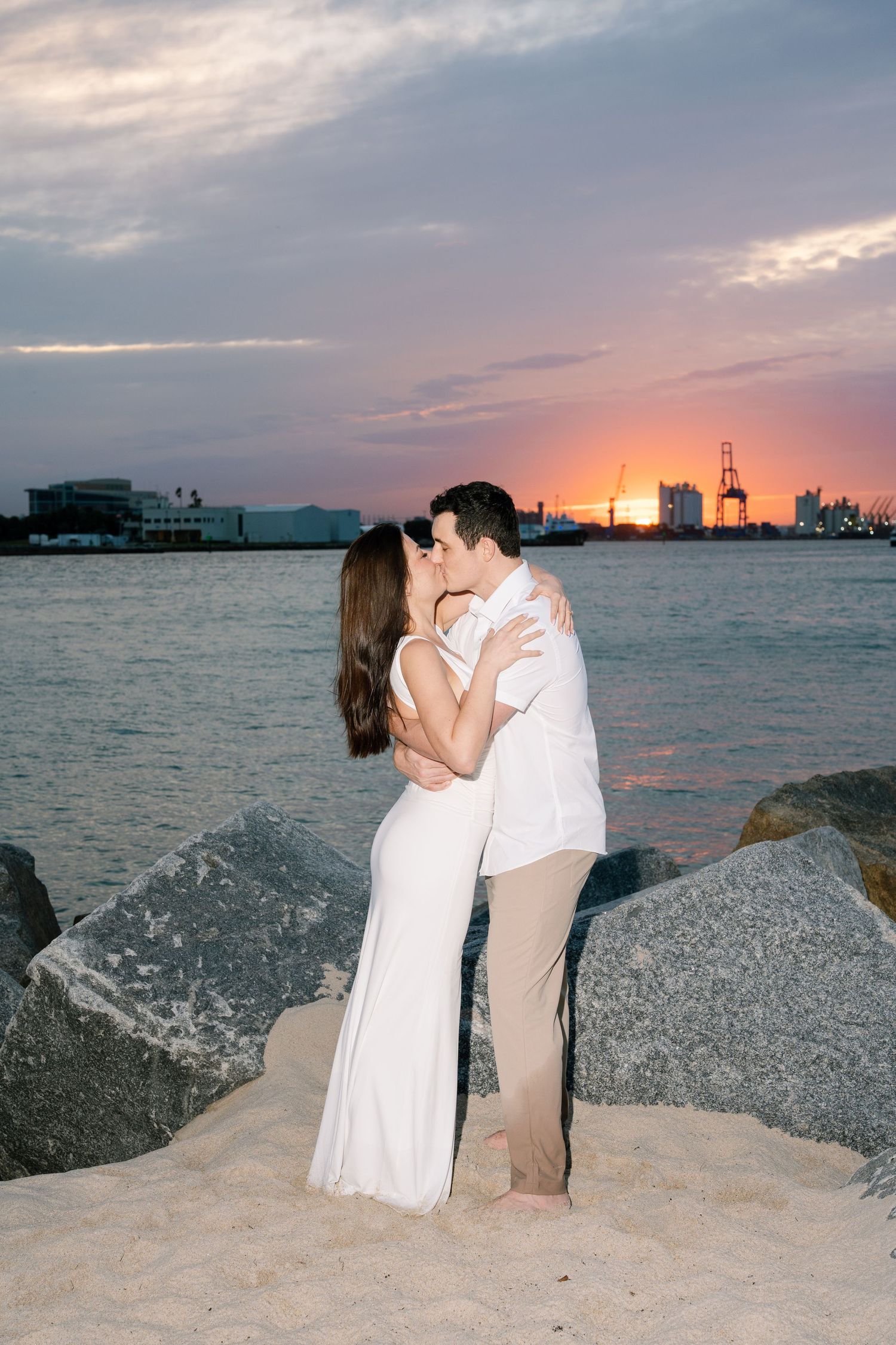 Couple embracing on rocky shore during sunset with ocean and city skyline in background.