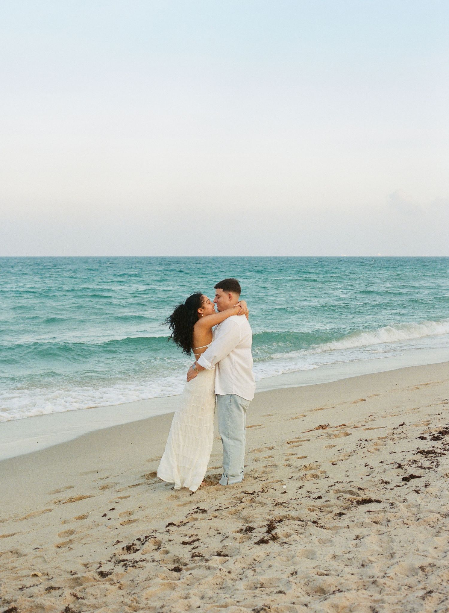 A couple in white attire shares a romantic moment on a serene beach with turquoise waters and soft sand.