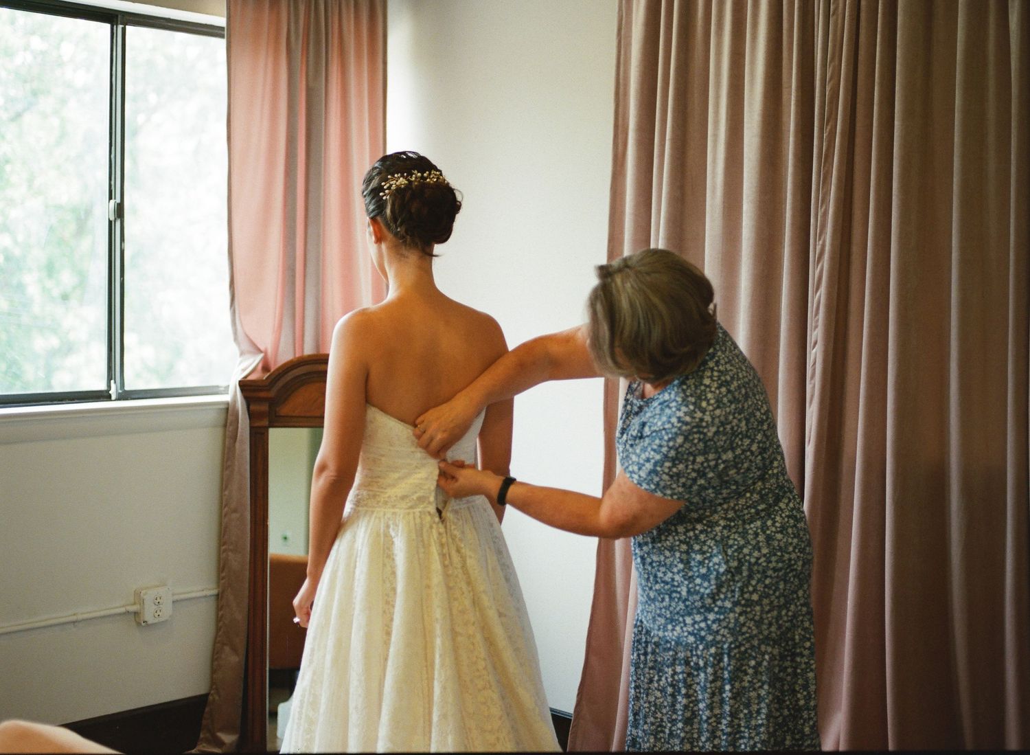 A bride and companion walk down a hallway with pink curtains during wedding preparations.