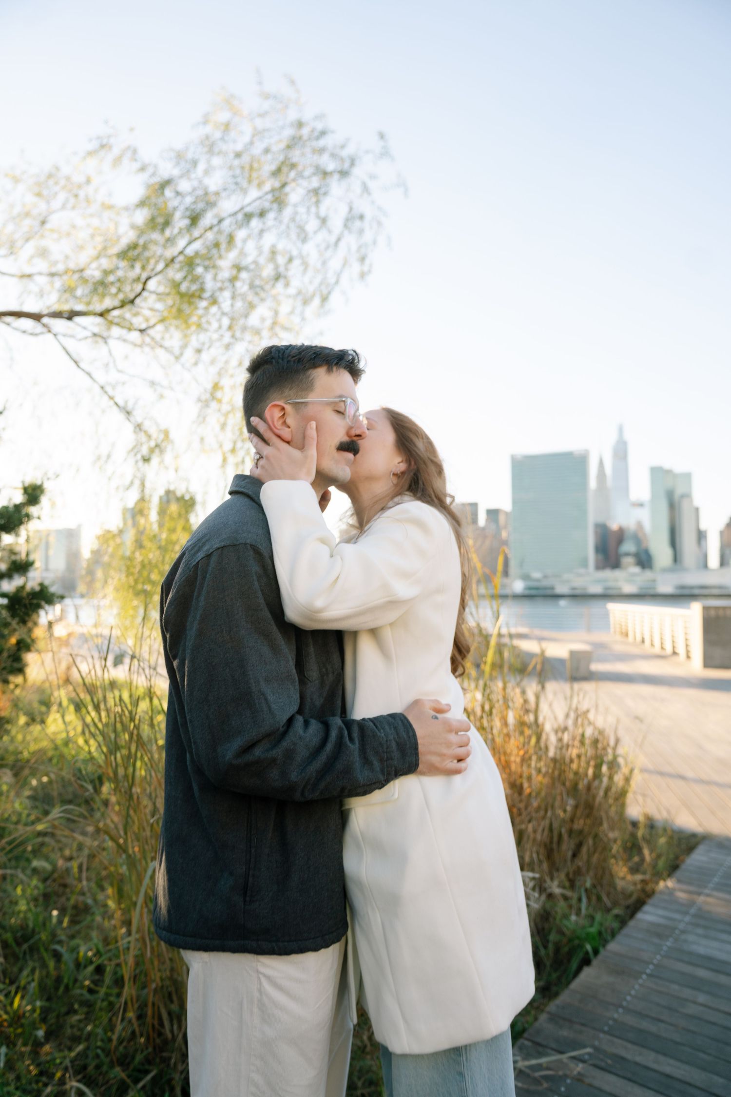 Sweet embrace shared on waterfront deck with iconic NYC buildings visible in distance.