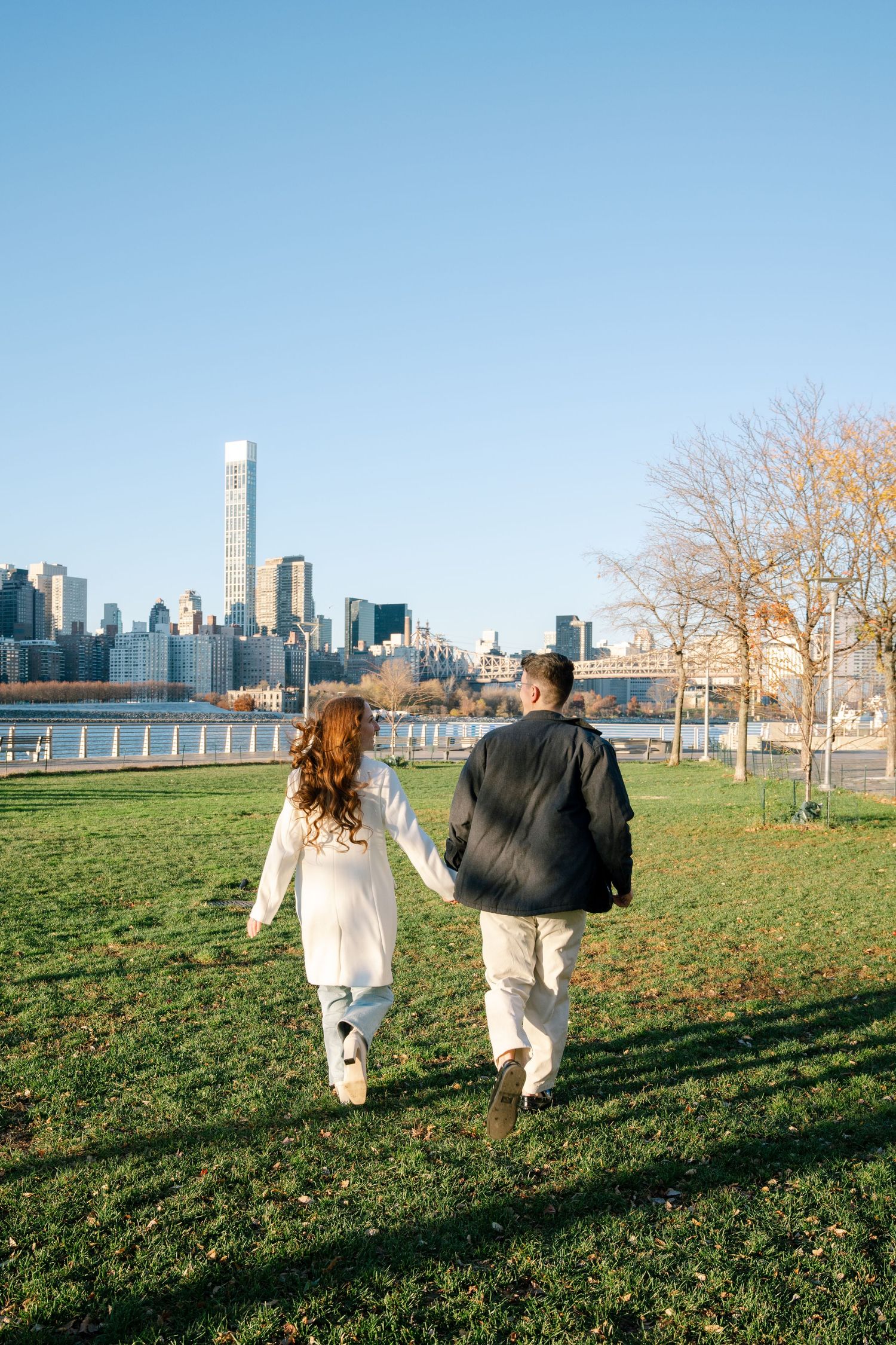 A couple walks hand in hand across a grassy field with the New York City skyline in the background.