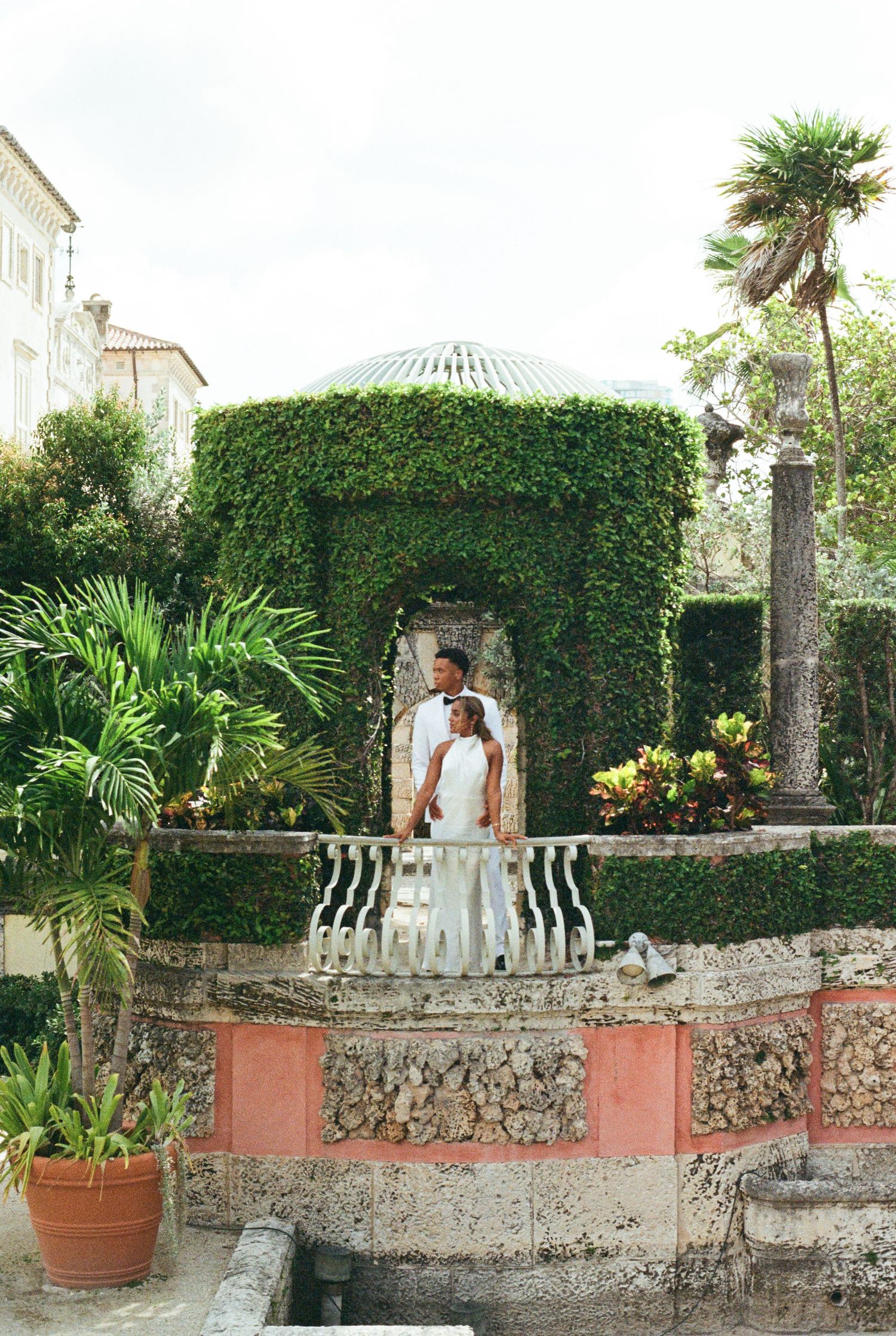 A panoramic view of a Mediterranean garden with stone walls, potted plants, and white chairs against a lush green hedge.