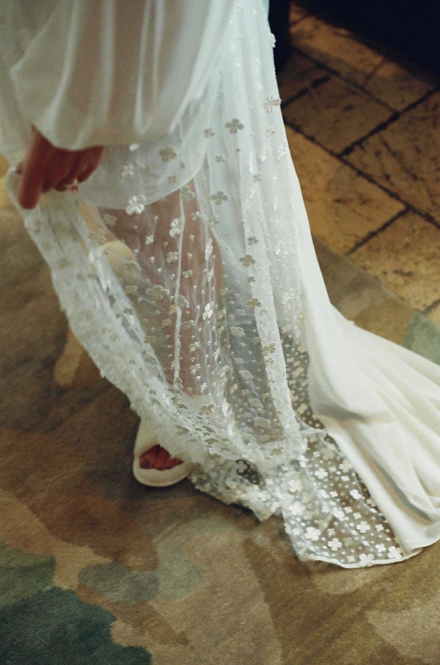 Close-up detail of intricate lace wedding dress hem trailing on stone floor.