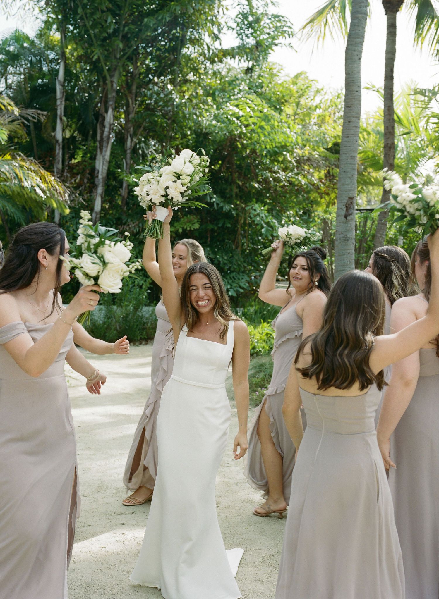 Smiling bride in white gown surrounded by bridesmaids in taupe dresses raising white floral bouquets in tropical setting.