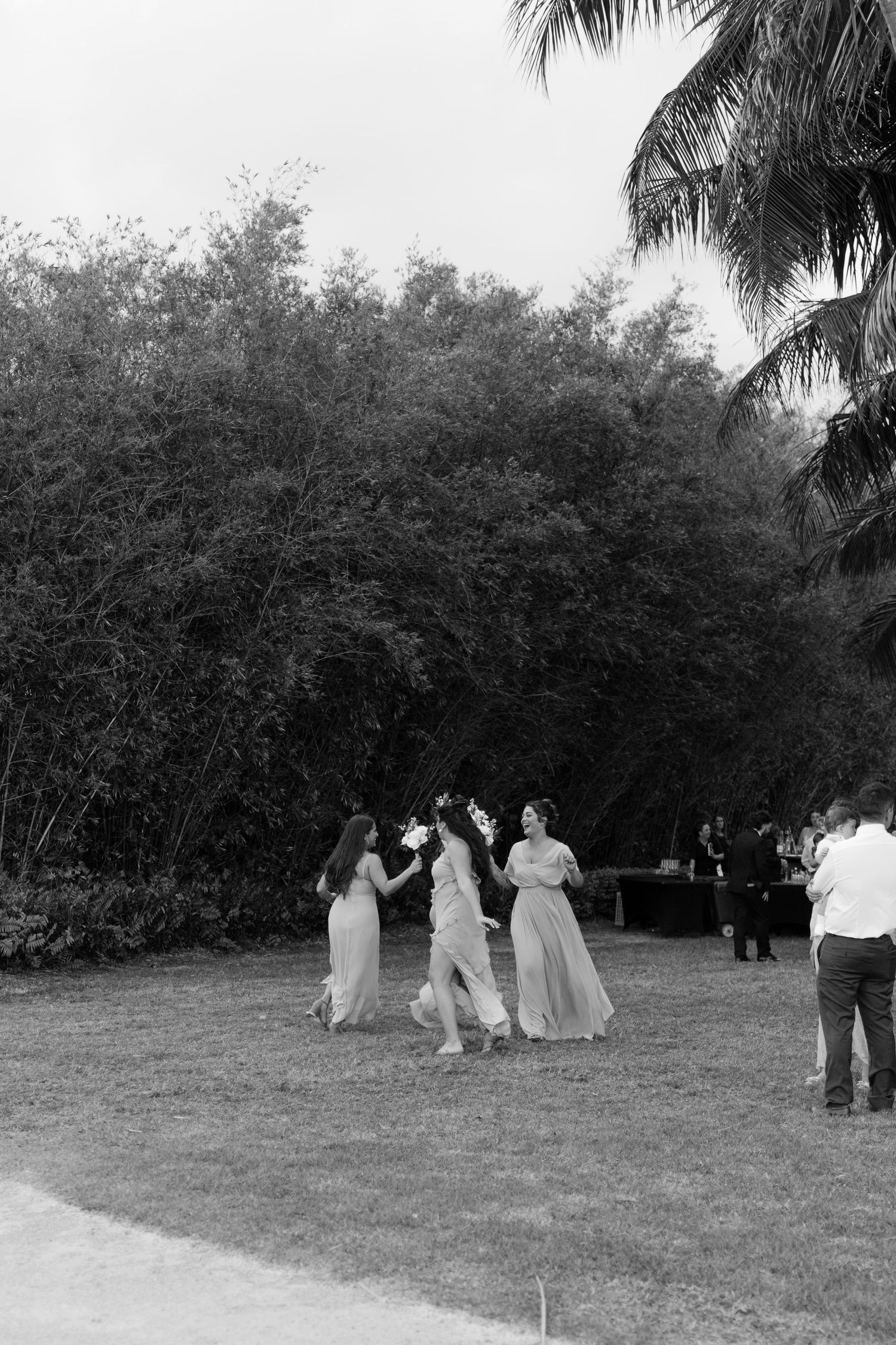 Bridesmaids in flowing gowns dancing joyfully on grass near tropical trees, black and white film photo.