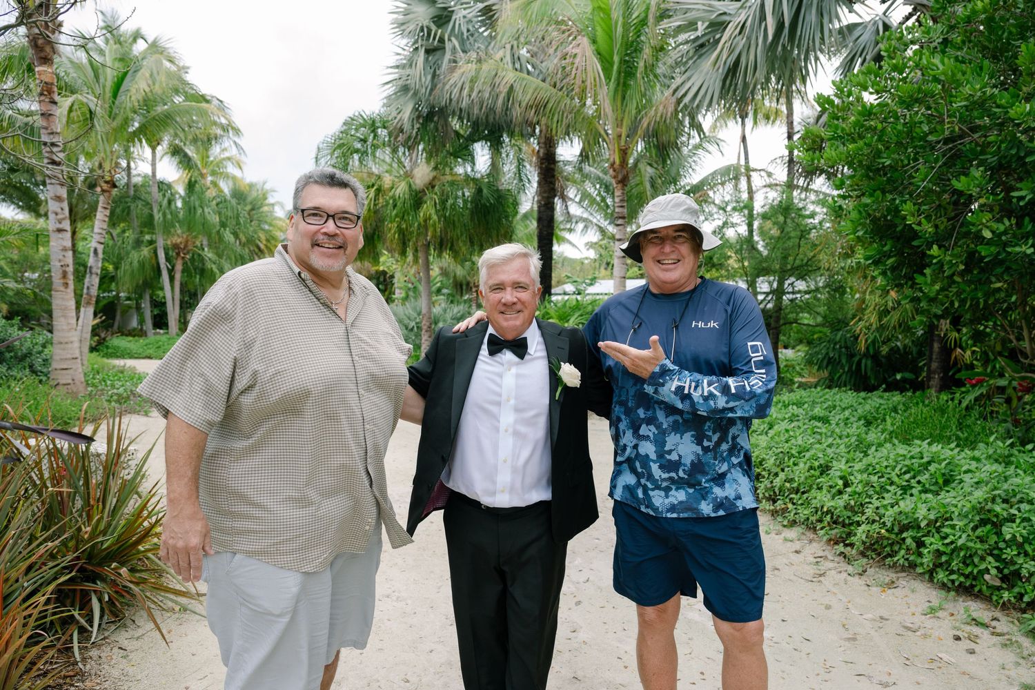 Three men smile together outdoors on a tropical path, one wearing a tuxedo with a boutonniere.