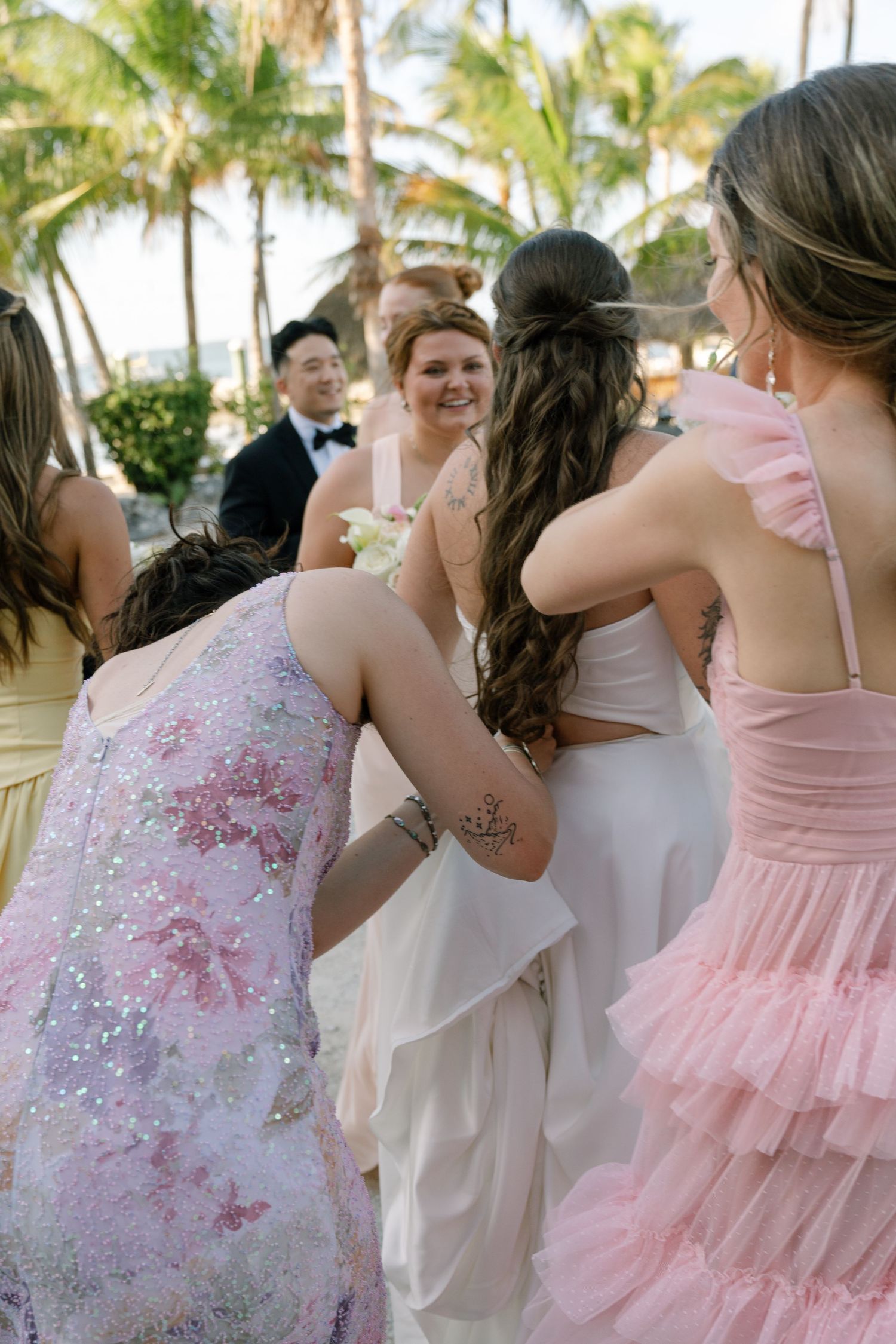 Bridesmaids in pink dresses gather around a bride in white at an outdoor tropical wedding ceremony with palm trees.