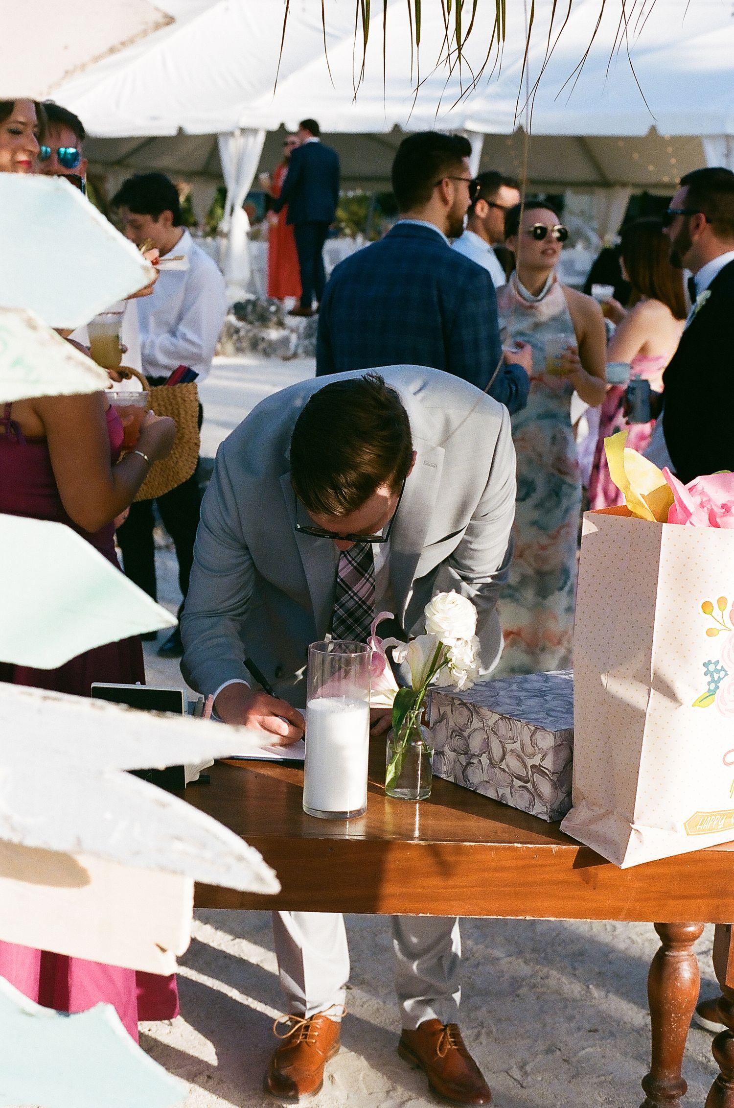 Groom in gray suit leaning over wooden table with flowers and gifts at outdoor beach wedding reception.