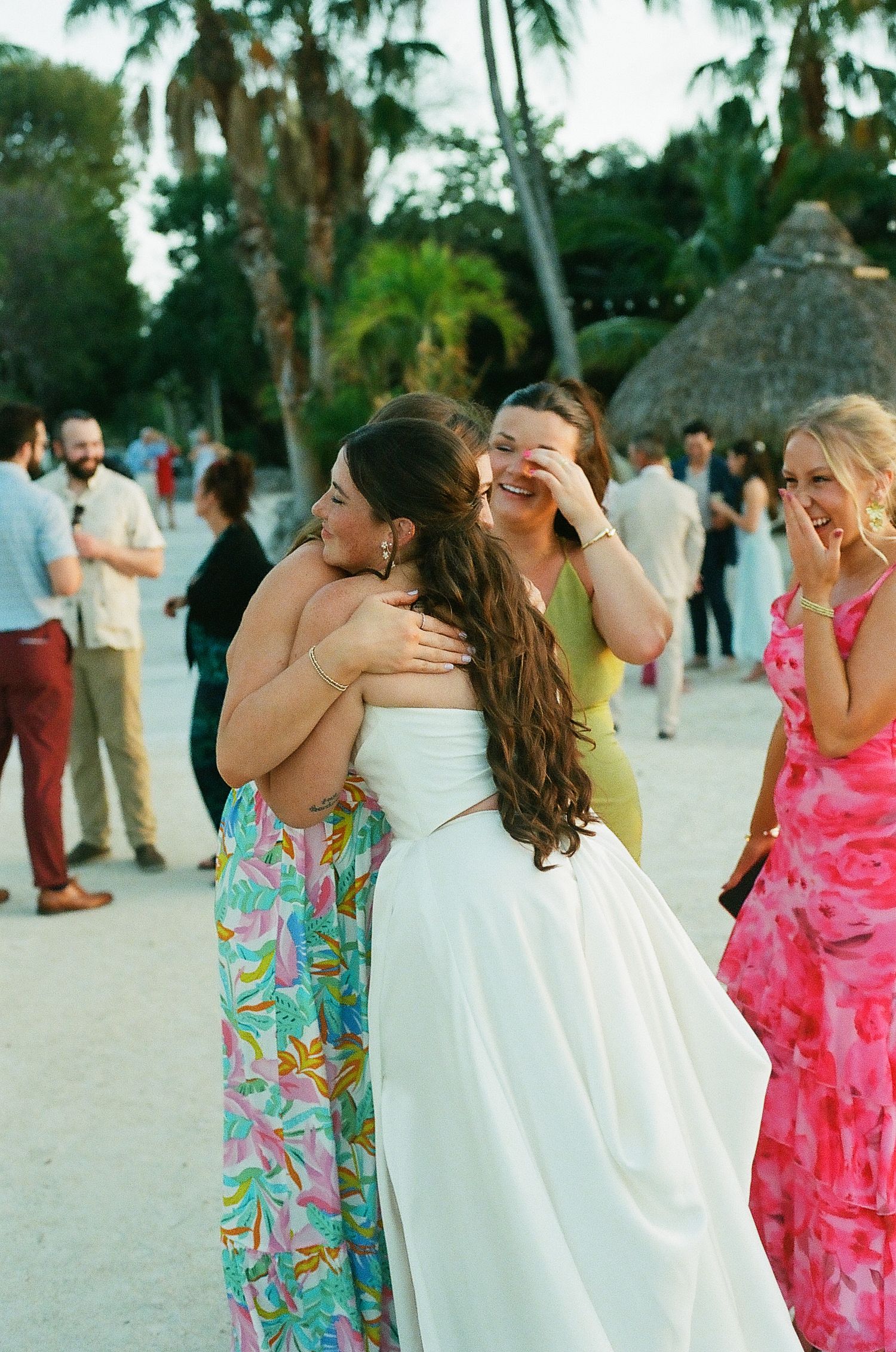 Bride in white gown receives warm hugs from guests in colorful dresses at an outdoor tropical beach wedding celebration.