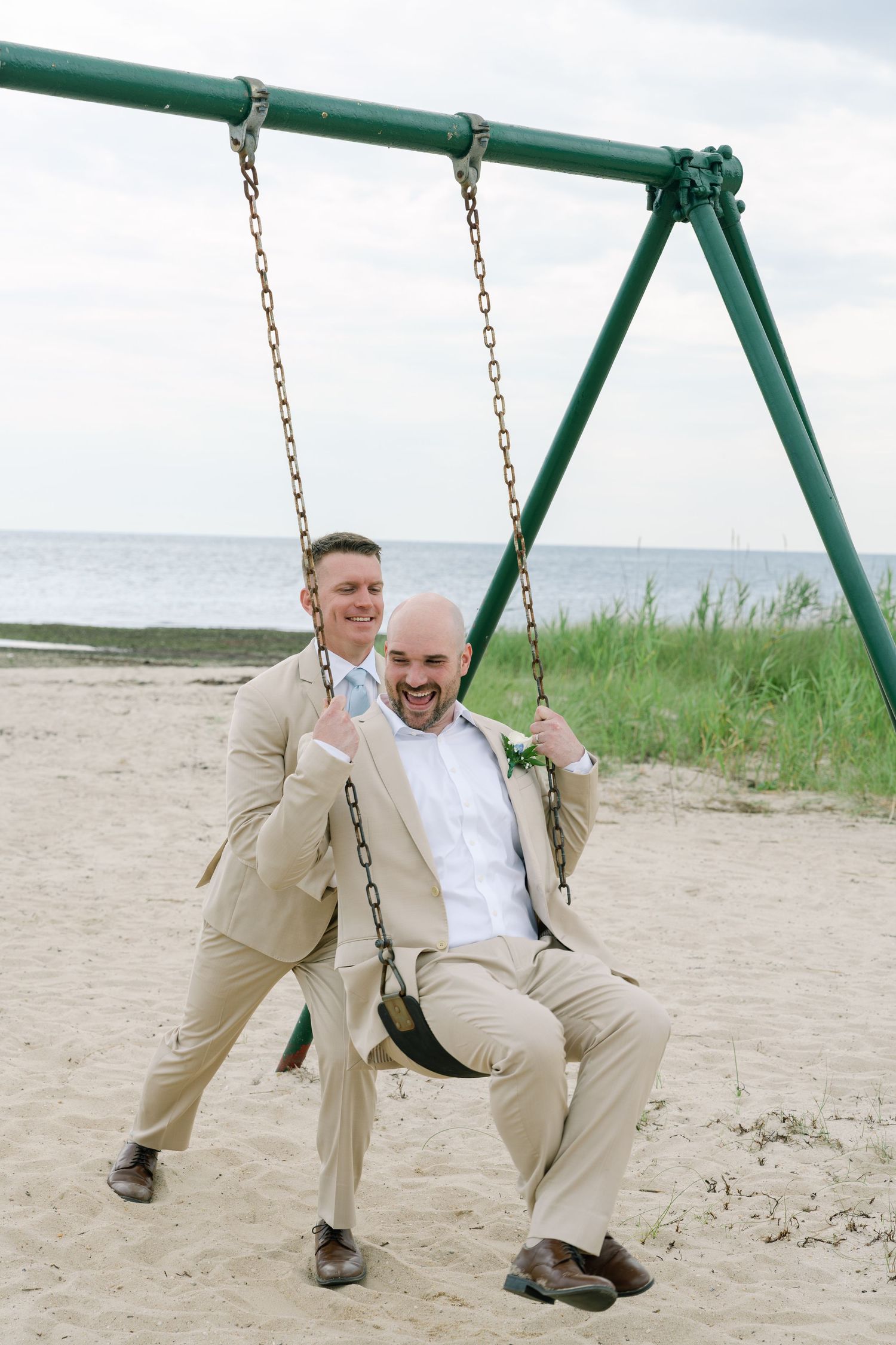Two groomsmen in tan suits laugh and play on a beach swing set near the ocean.