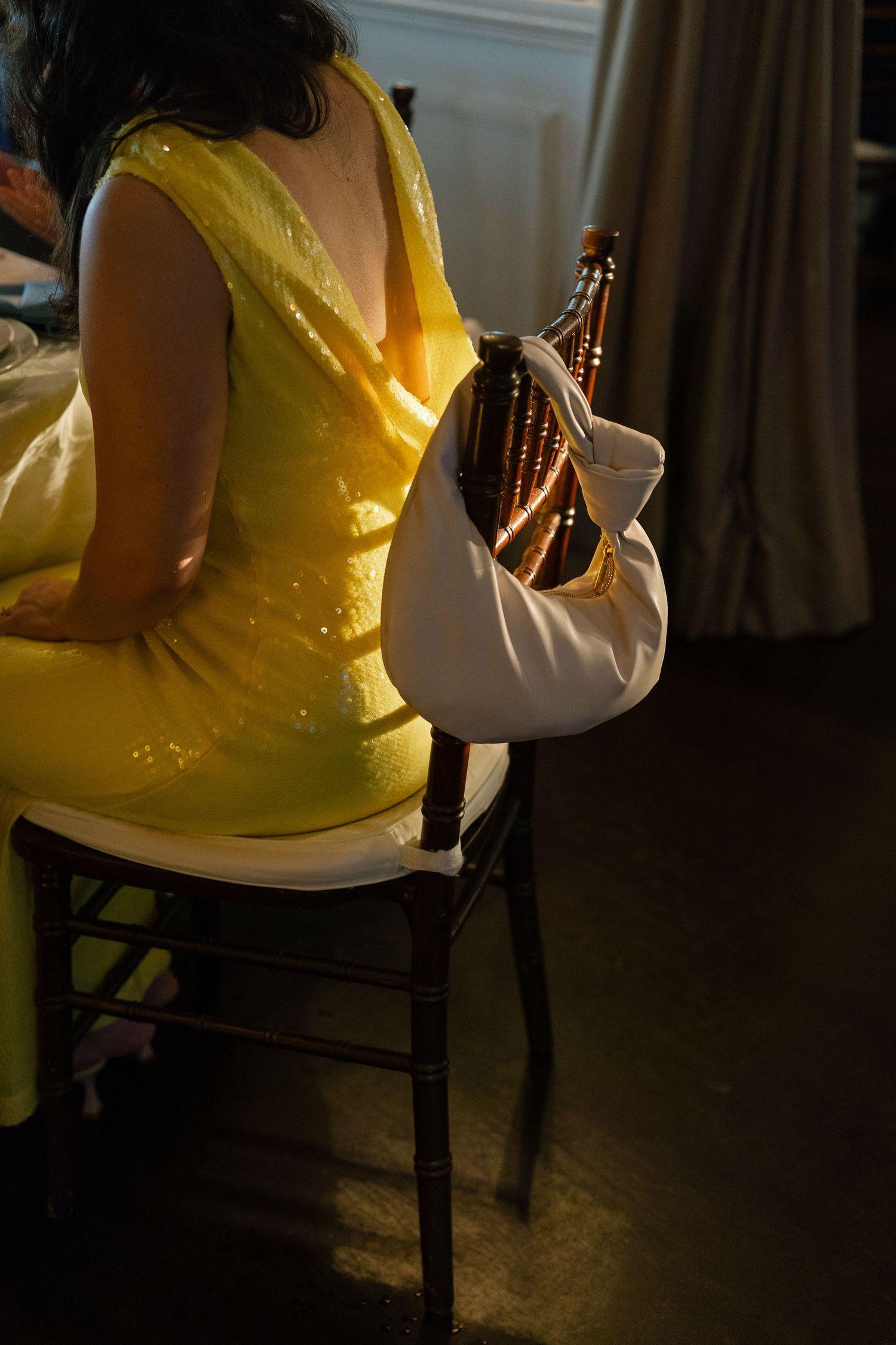 Woman in yellow sequin dress seated on chair holding white bag in warm light.