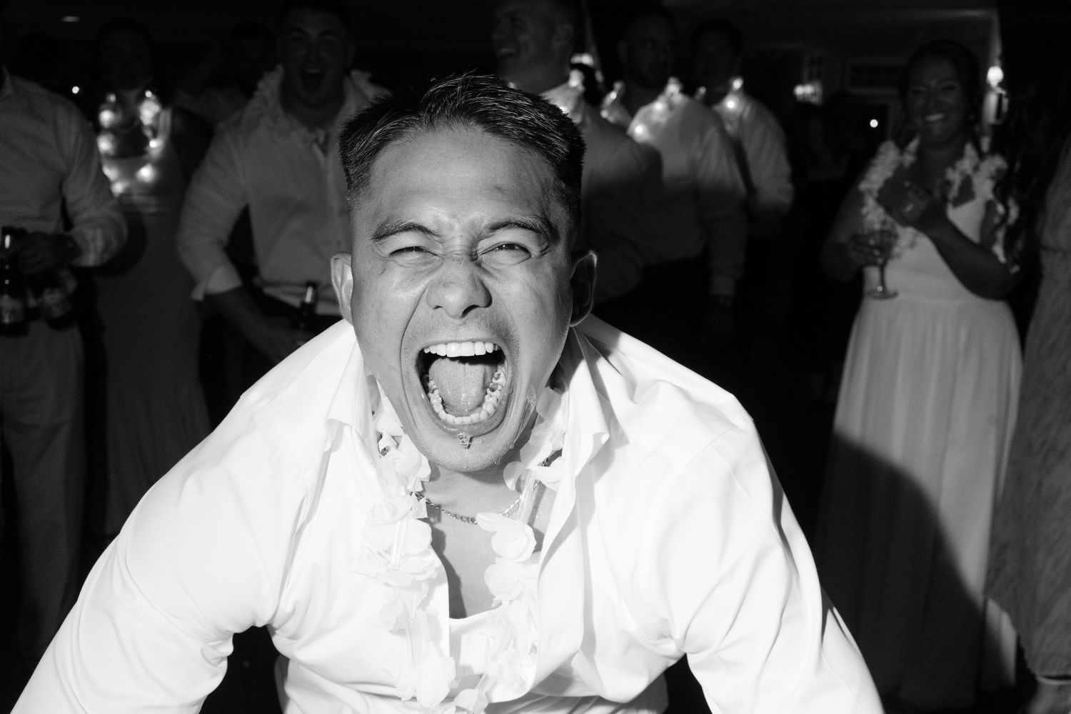 Black and white photo of a man laughing with his mouth wide open at a festive wedding reception.