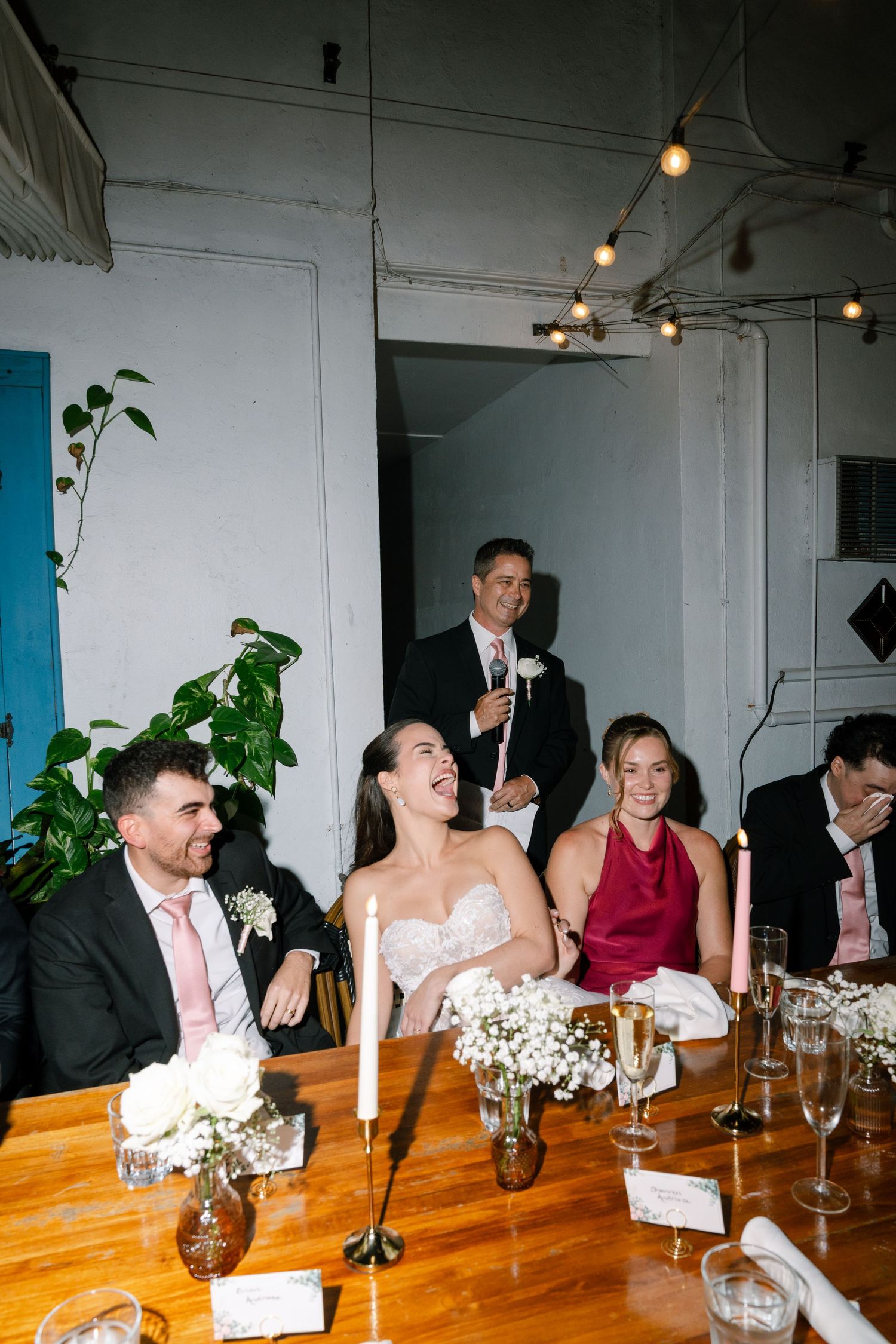 Bride laughs joyfully at wedding reception dinner table during heartfelt speech by standing guest.