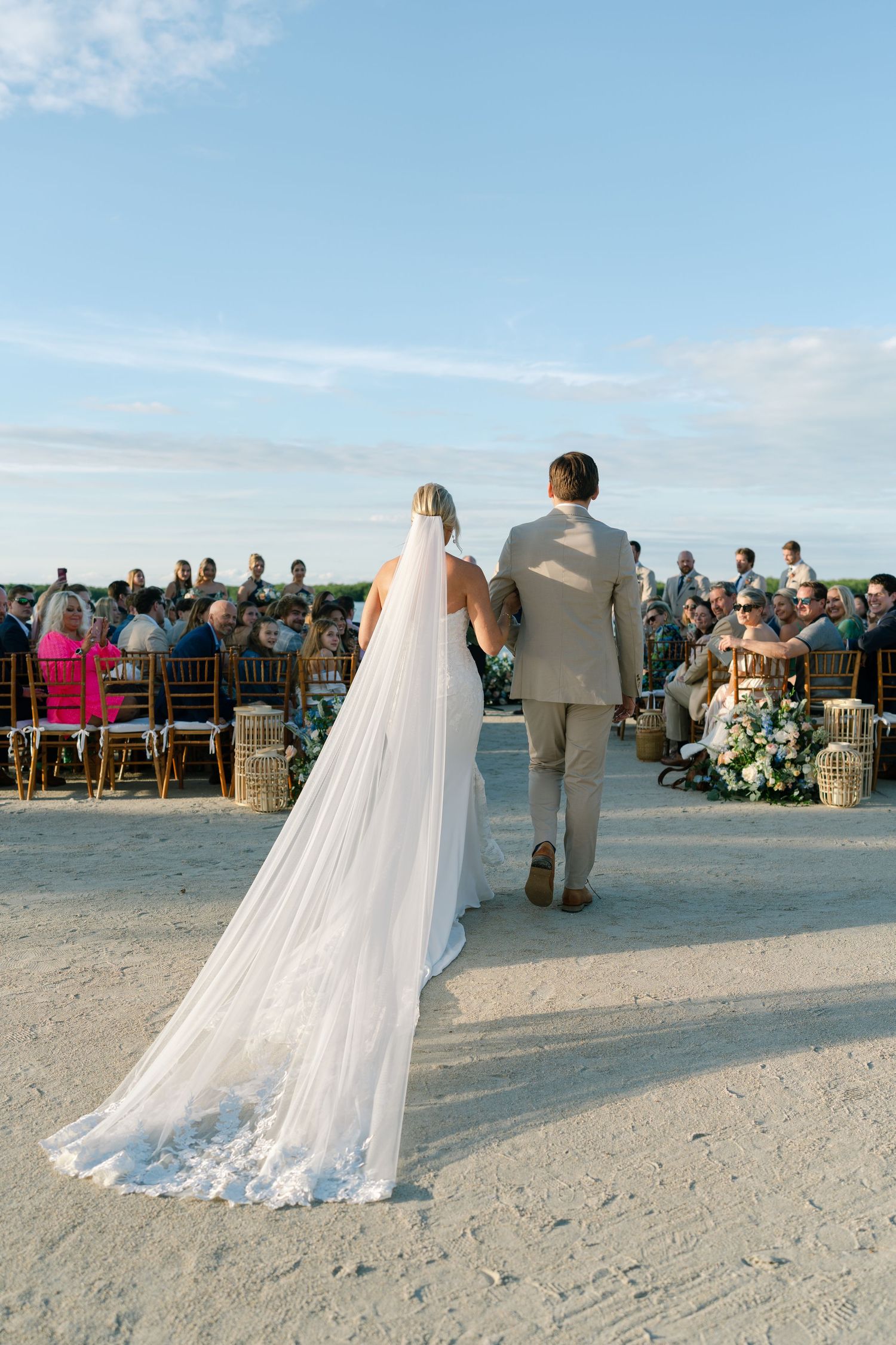A beachside wedding ceremony with guests seated in wooden chairs as the couple exchanges vows against an ocean backdrop.