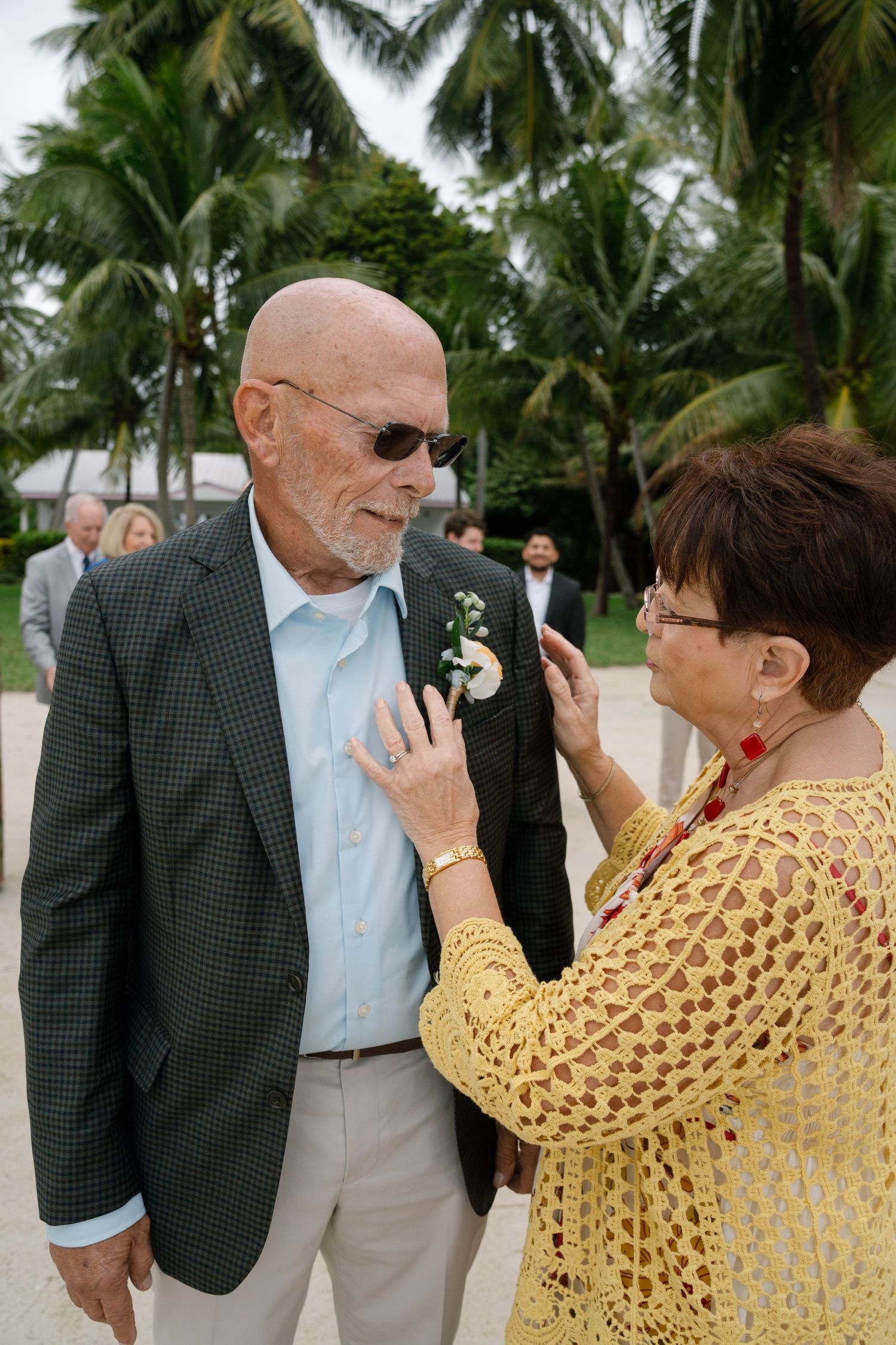 A woman in a yellow crochet shawl interacts with a man wearing sunglasses and a suit jacket outdoors among palm trees.