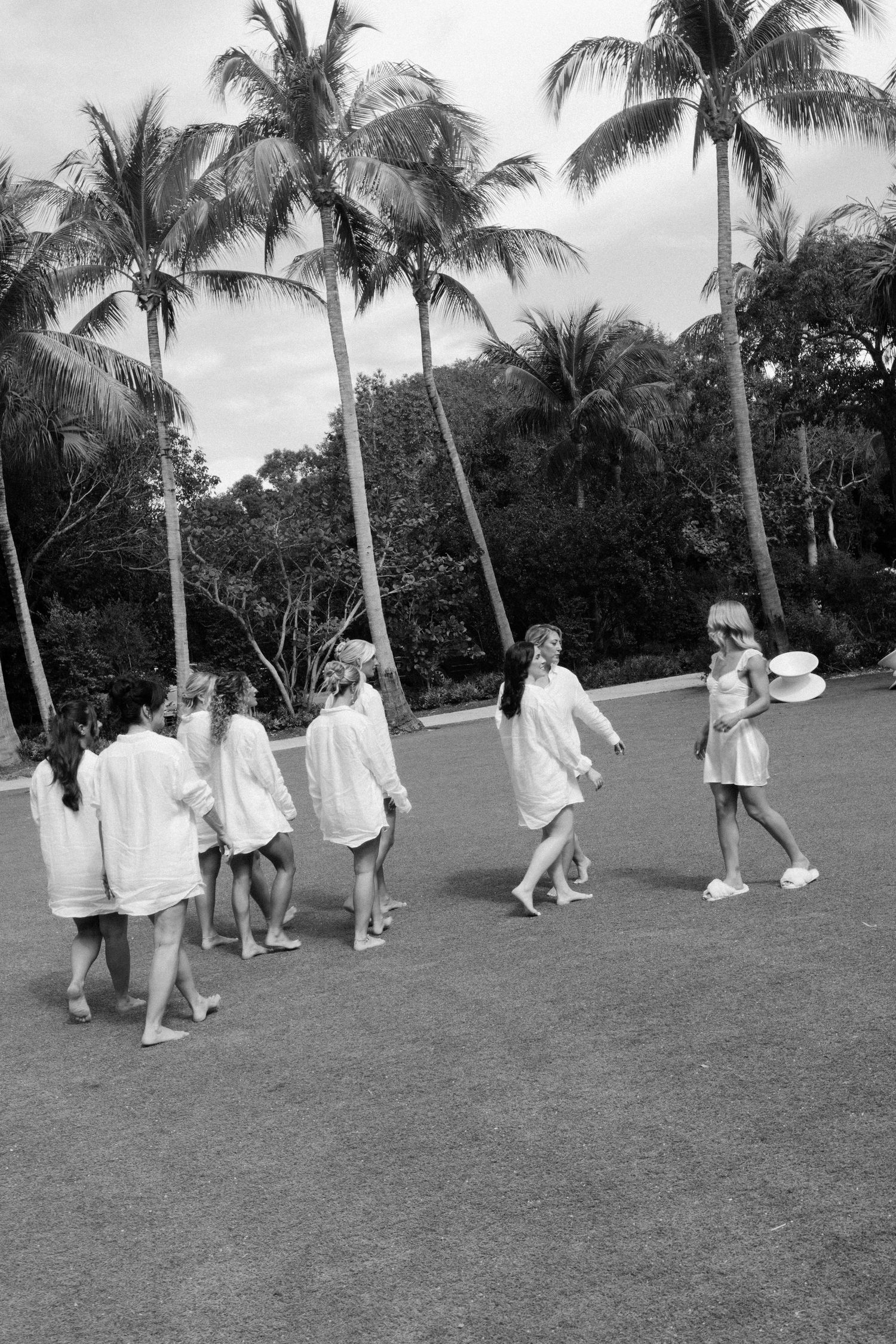 A line of people in white attire walk across a palm tree-lined lawn during an outdoor gathering.