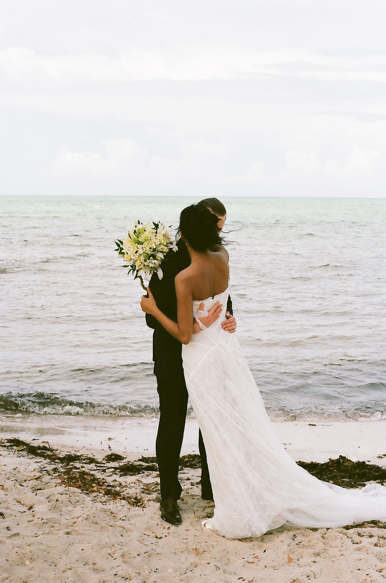 Bride and groom embrace on a sandy beach, with the bride holding a white bouquet and wearing a lace wedding gown.