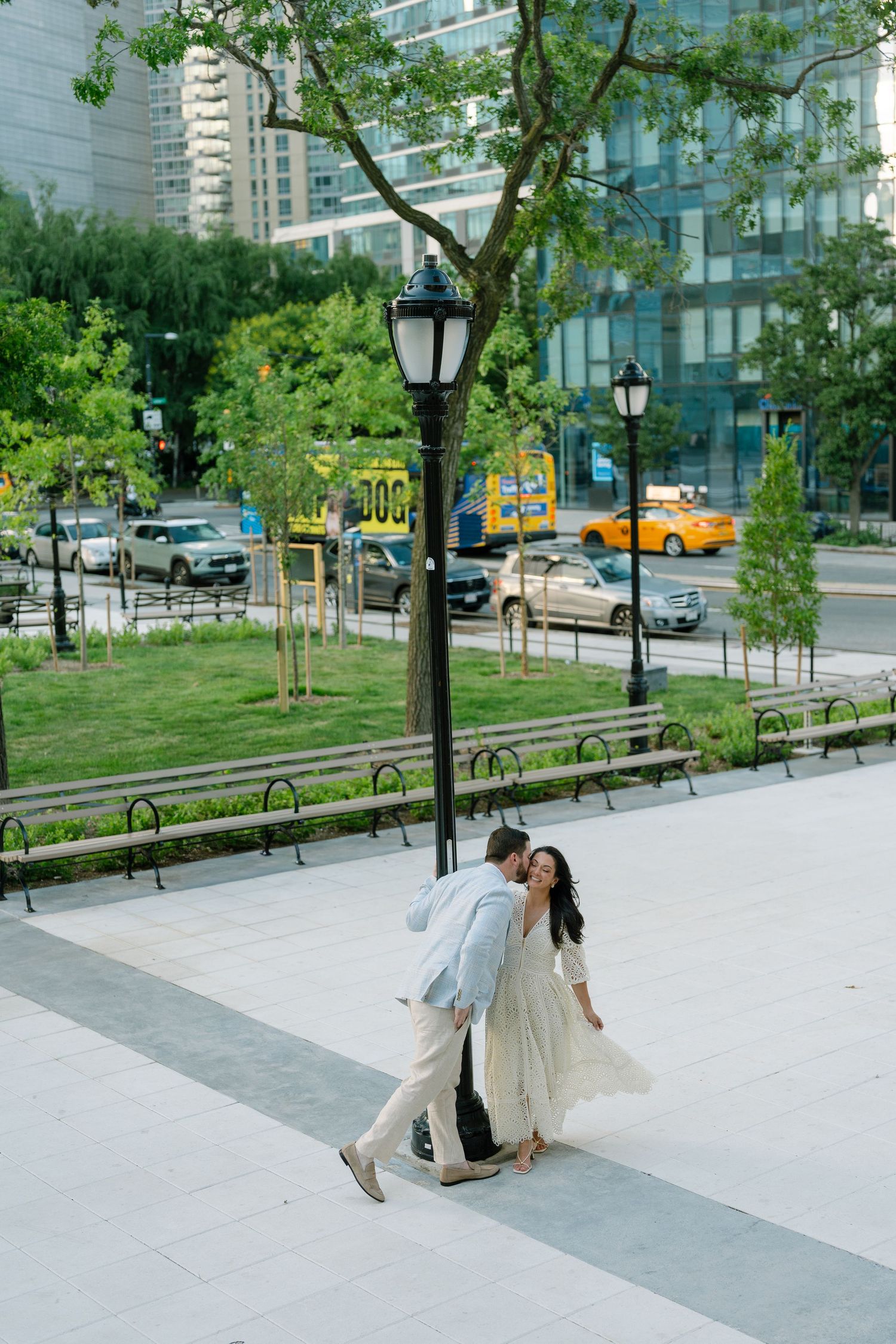 Couple sharing a romantic dip kiss on a city plaza sidewalk near a lamppost, with urban buildings and traffic in the background.
