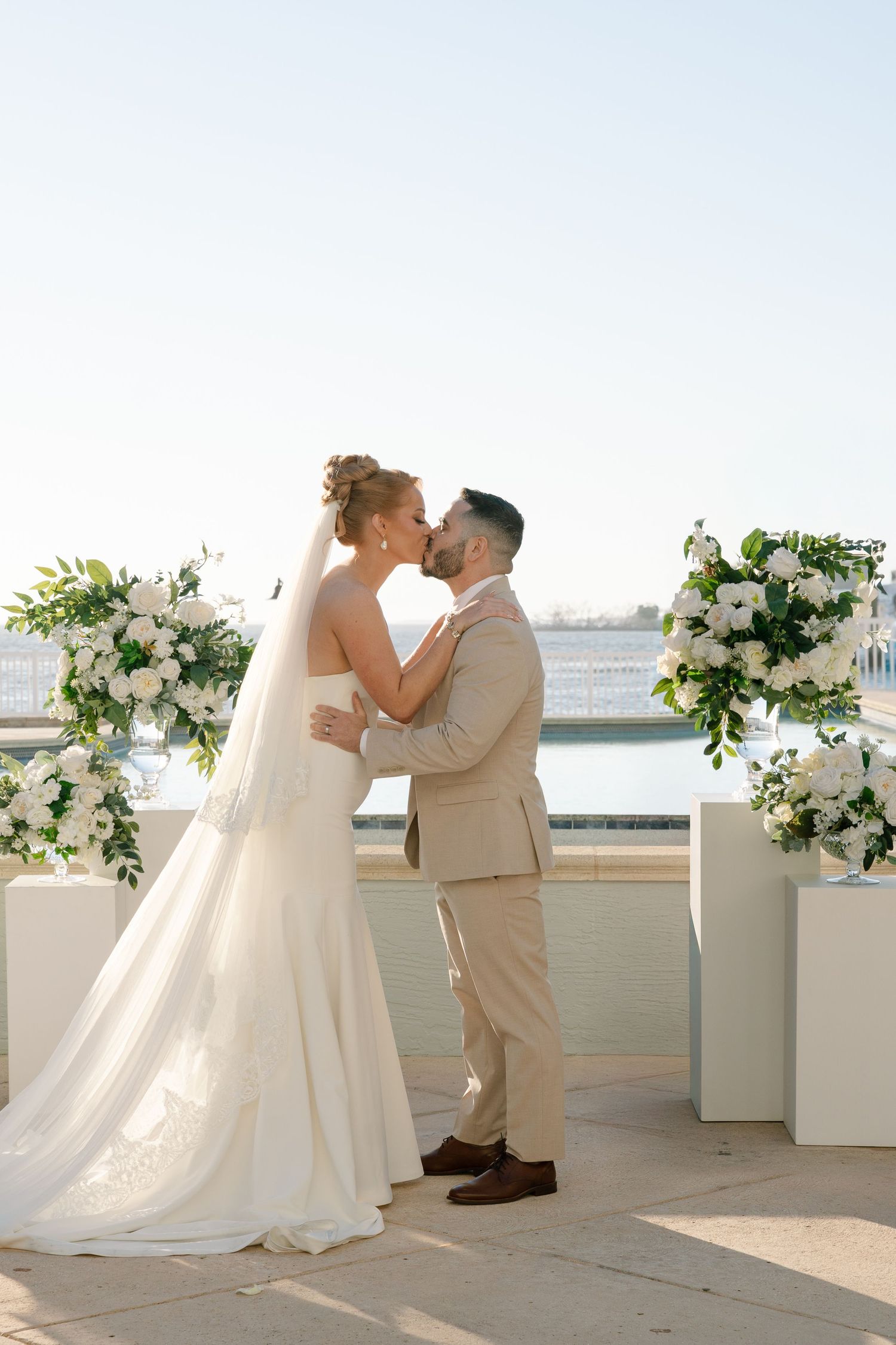 A romantic wedding couple shares a tender moment on a white-decorated terrace overlooking the water.