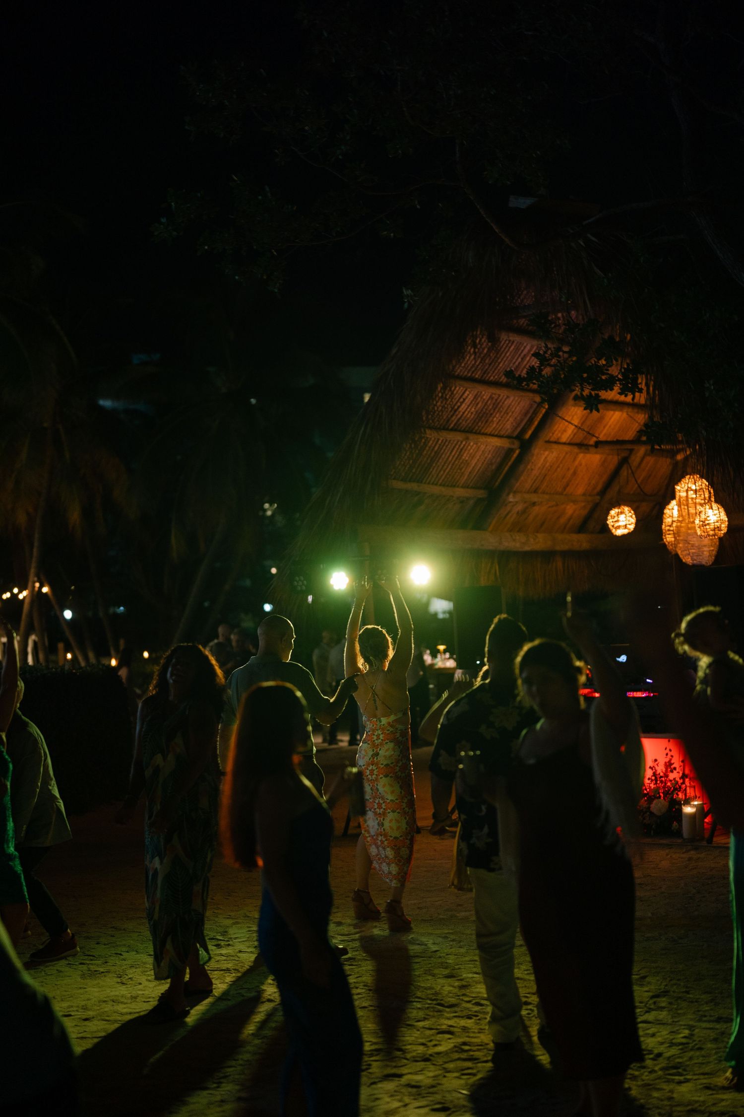 People dancing at a nighttime beach party under a thatched roof with glowing lanterns.