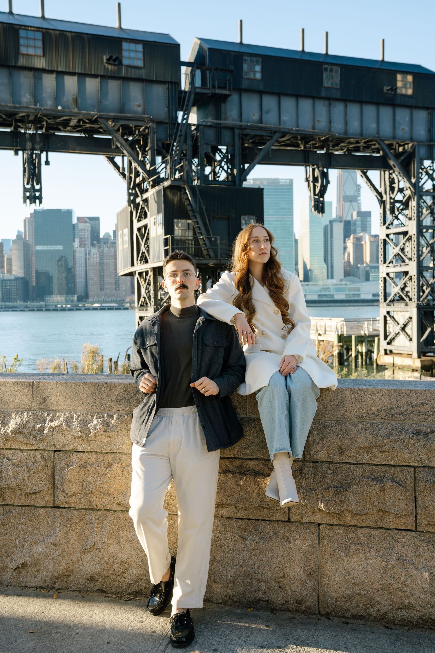 Couple in coordinated white outfits posing against urban waterfront backdrop with city skyline visible.