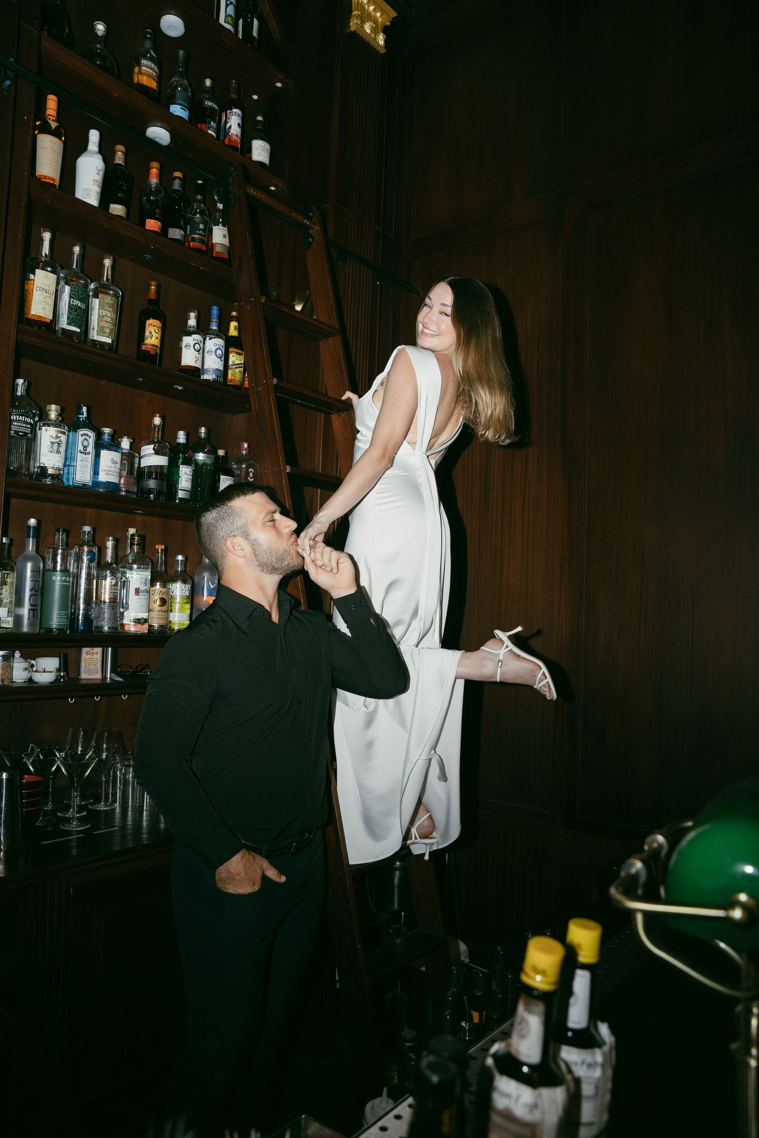 Person in a black outfit supporting another in a white dress at a dimly lit bar with shelves of bottles in the background.