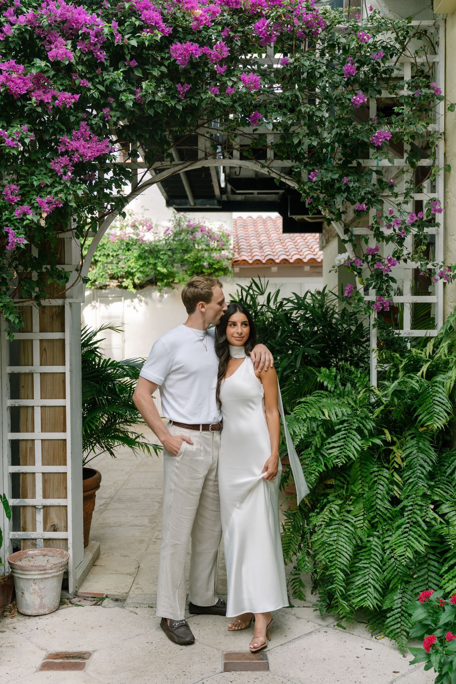 Couple embracing under a floral arch with purple bougainvillea, surrounded by lush greenery and white lattice.