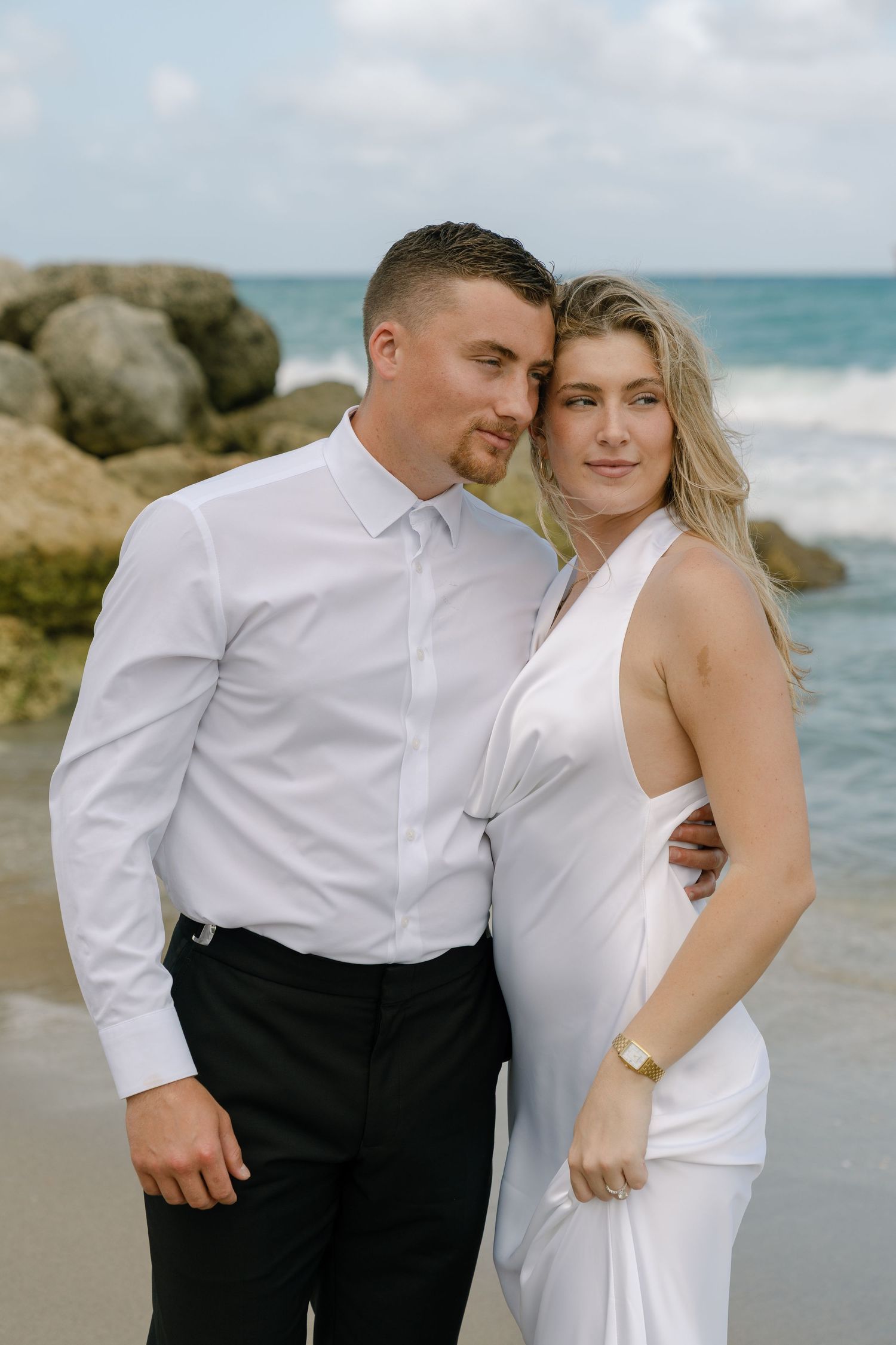 Couple in white attire posing on a rocky beach with ocean waves, woman in white dress and man in white shirt.