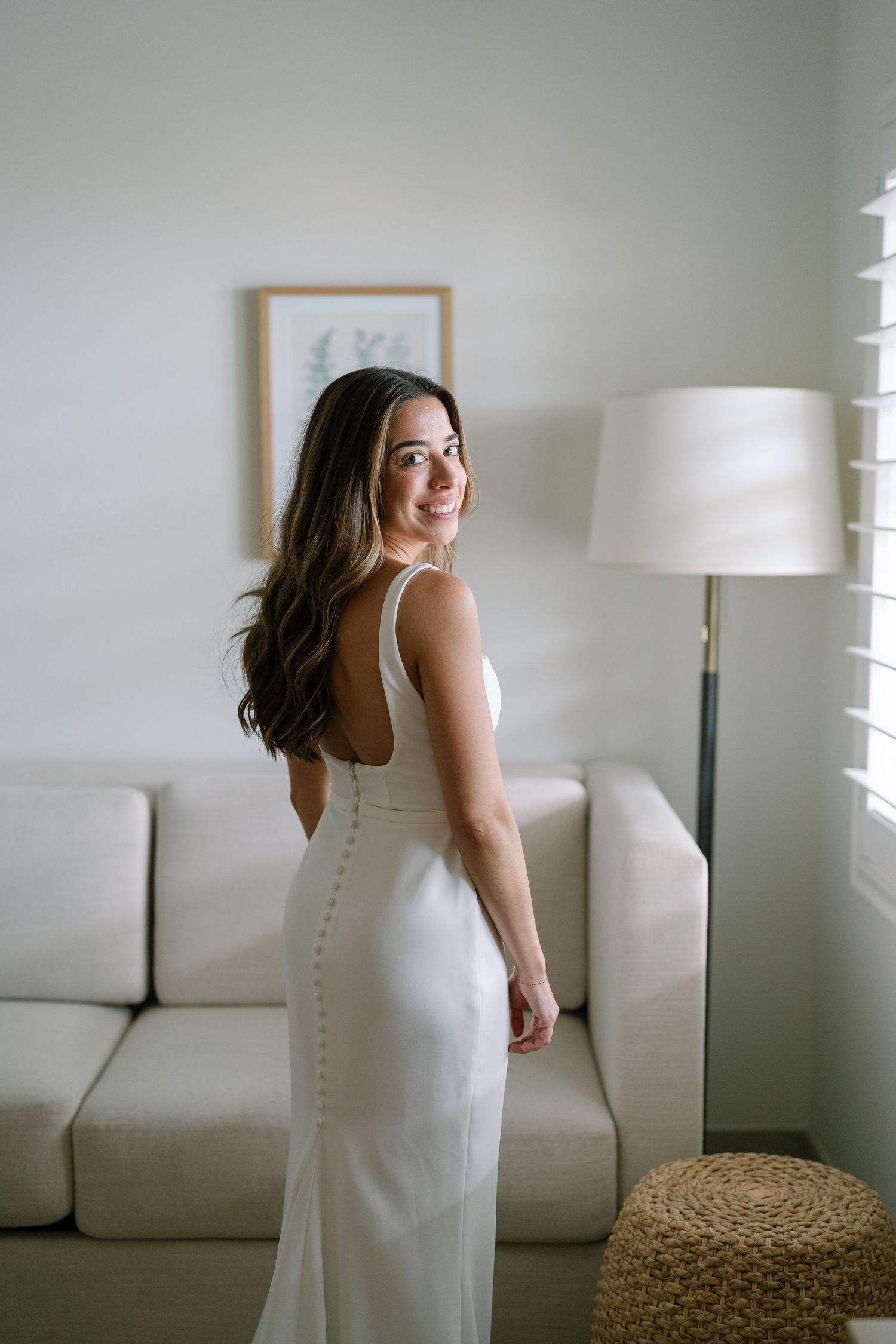 Bride in sleek white gown with button back detail smiling over shoulder in bright room.