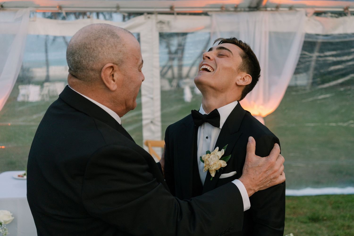 Emotional groom in black tuxedo sharing a heartfelt moment with an older man at an outdoor wedding reception.