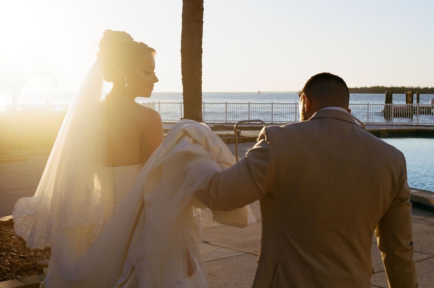 Two people walk along a scenic coastal boardwalk at sunset with sunlight streaming through the wooden railings.