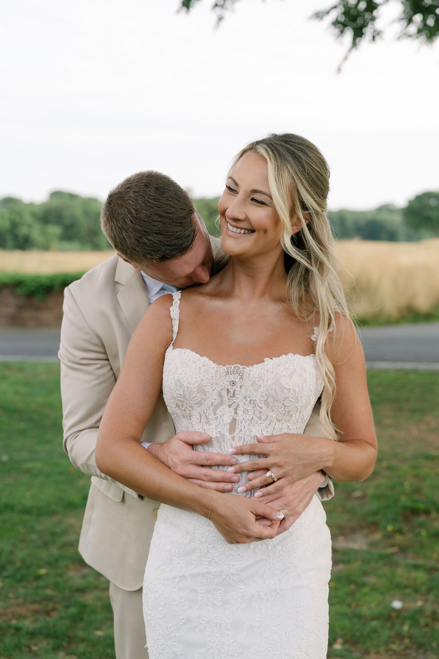Smiling bride in lace wedding gown embraced by groom in tan suit outdoors with golden fields in background.