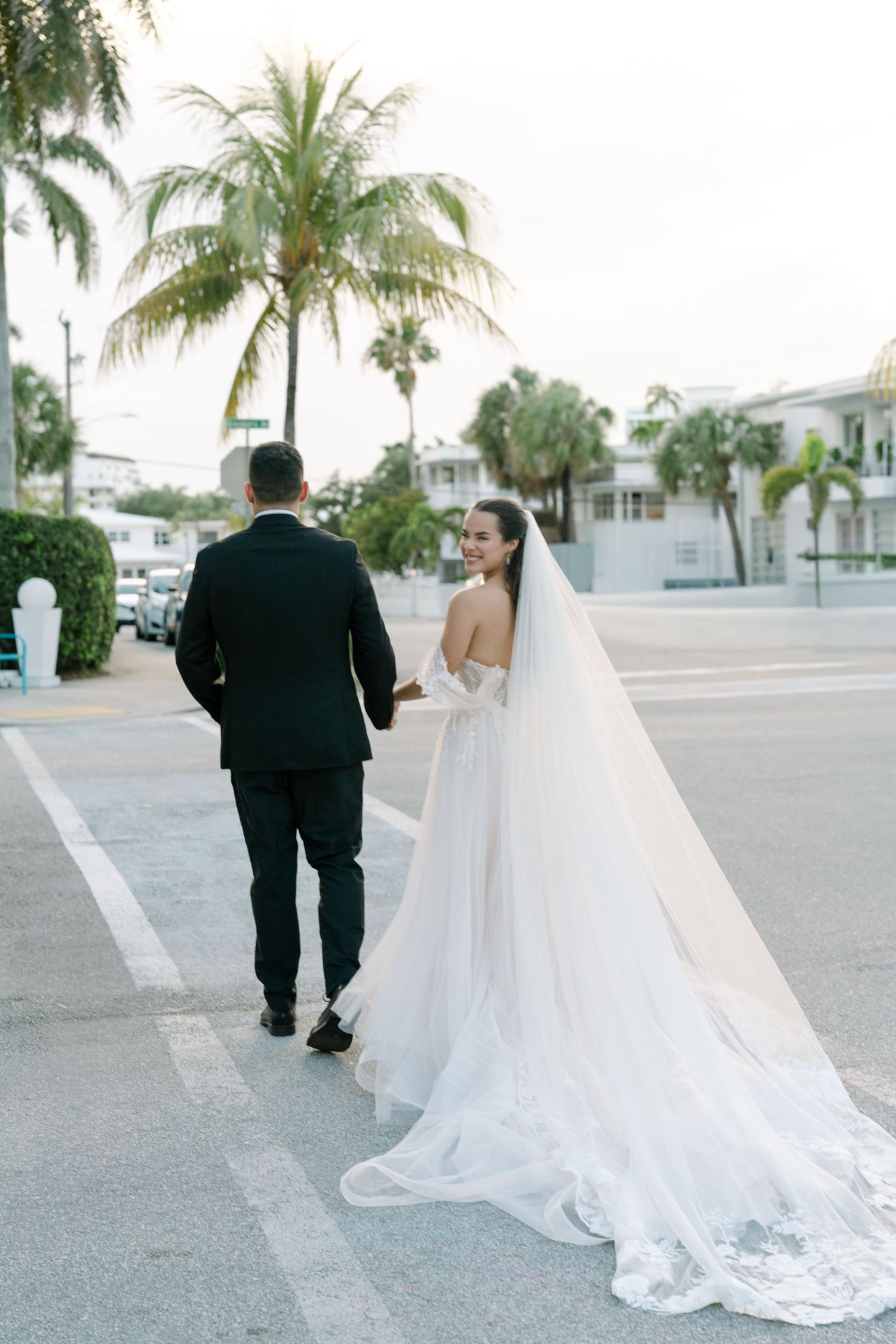 Bride in a flowing white gown and veil walks hand-in-hand with groom in black tuxedo on a sunny Florida street.