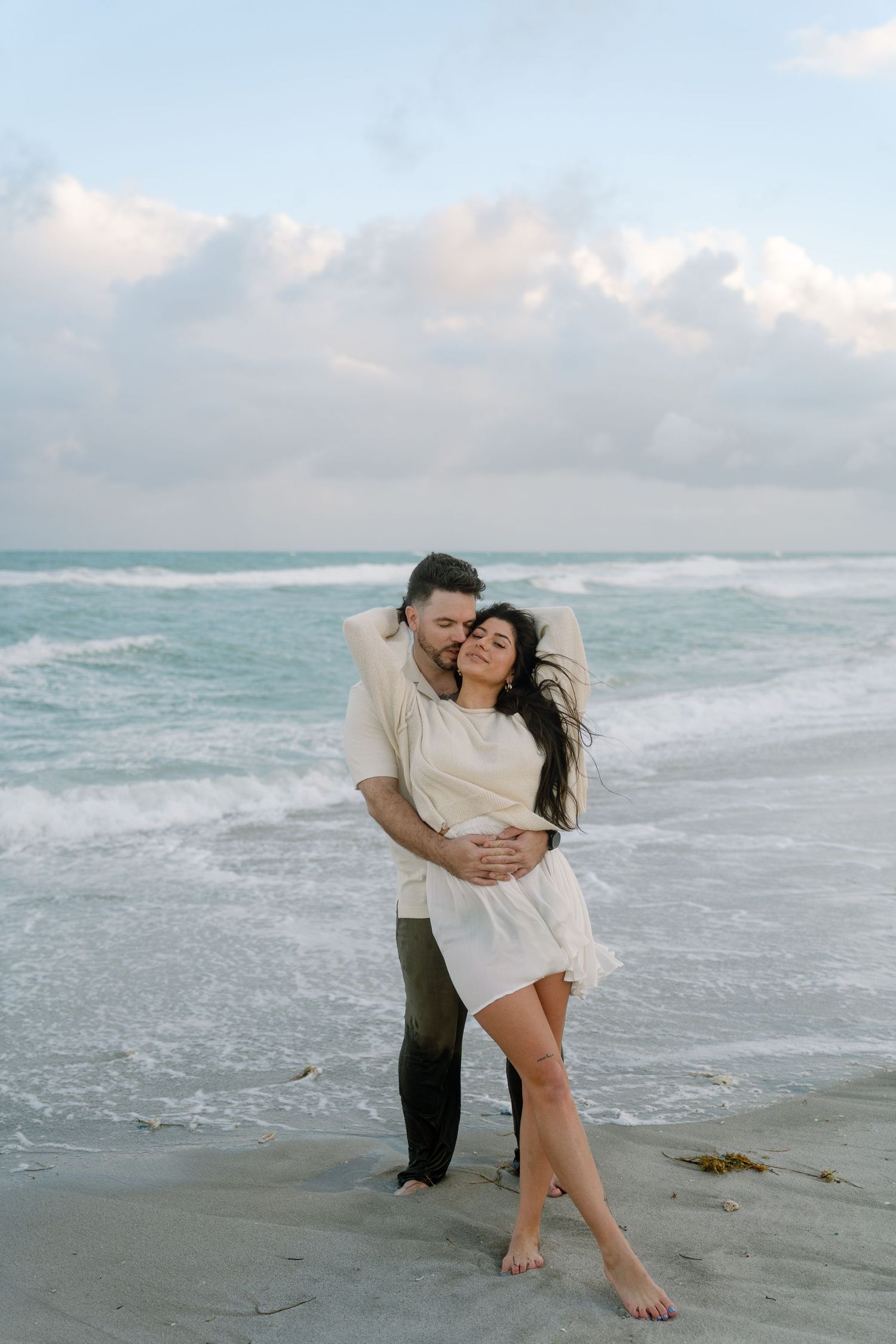 A couple embraces on a cloudy beach with crashing waves in the background, creating a romantic coastal scene.