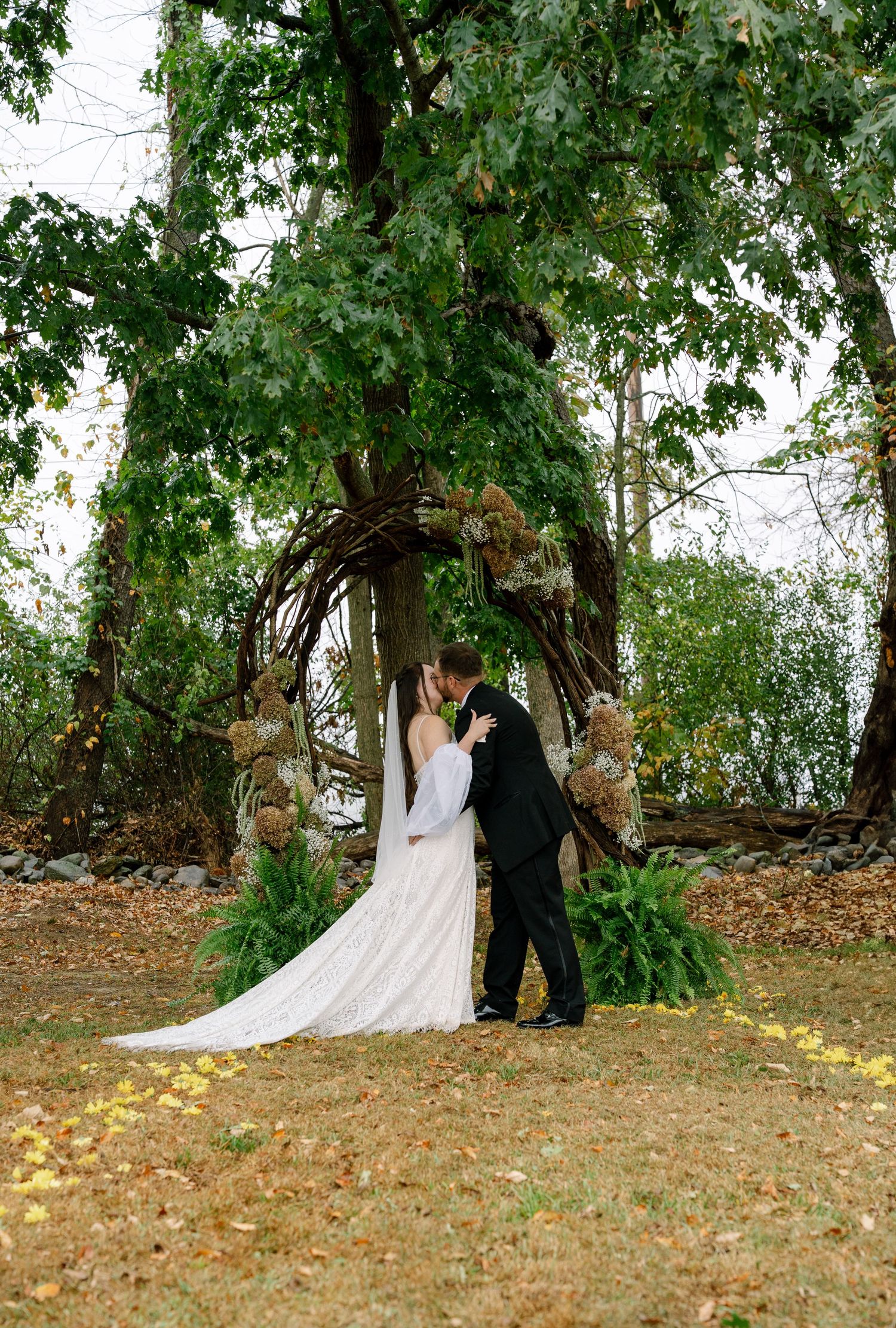 A couple shares a romantic moment on a hillside covered in fallen leaves beneath towering trees.