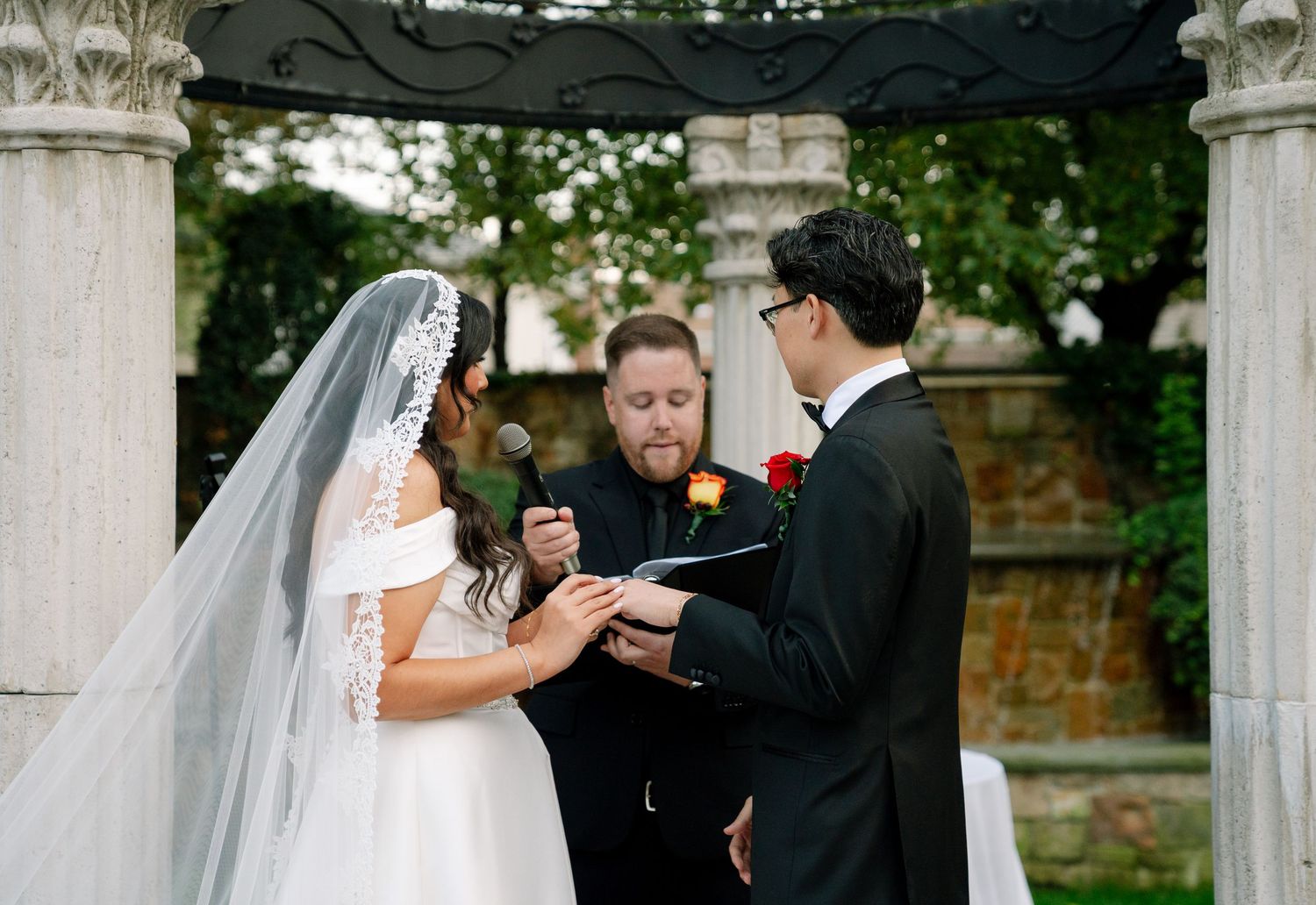 Outdoor wedding ceremony under white columns with couple exchanging vows.