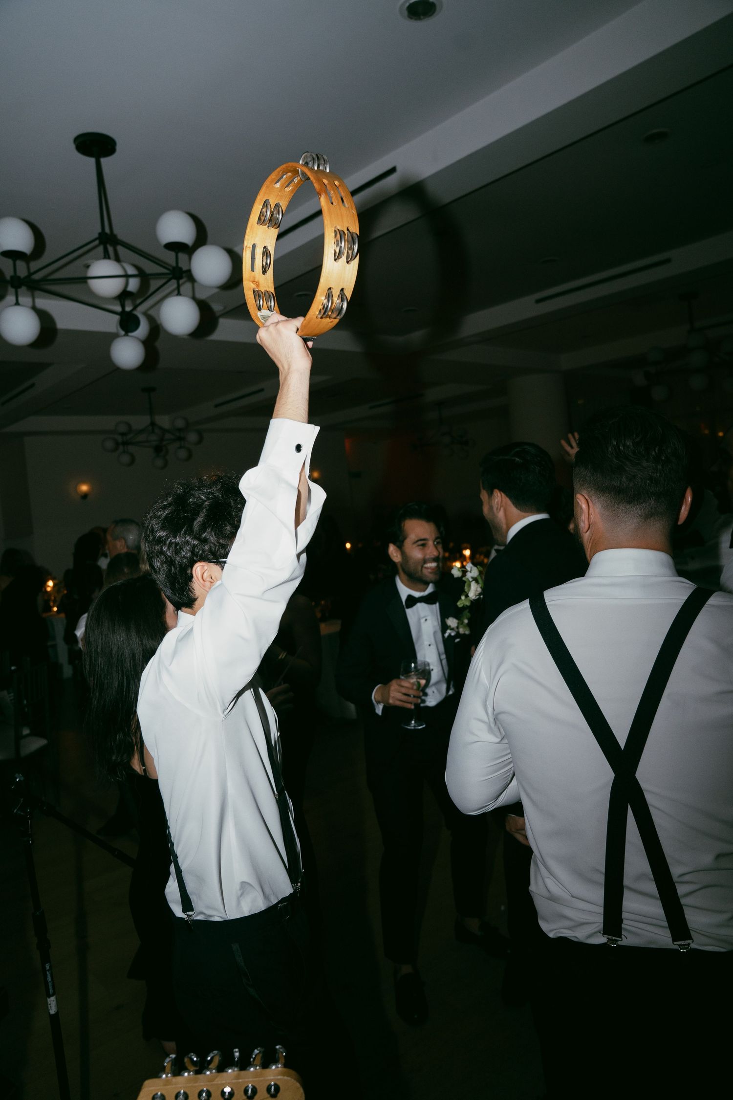 Someone holds up a gold tambourine on a dance floor at a formal event with partygoers in suits and suspenders.