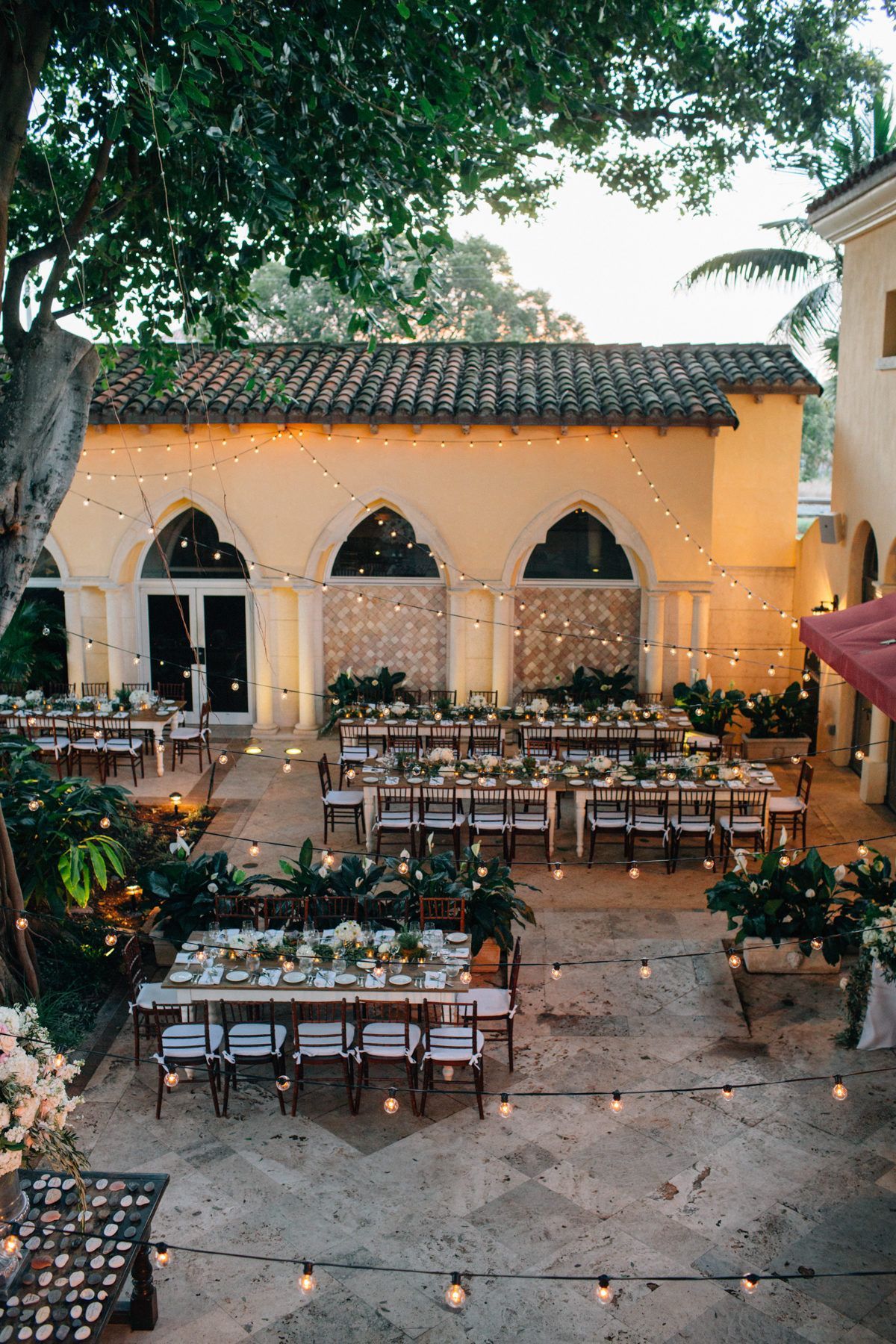 String lights illuminate an elegant outdoor wedding reception setup with long wooden tables and chairs in a Mediterranean courtyard.