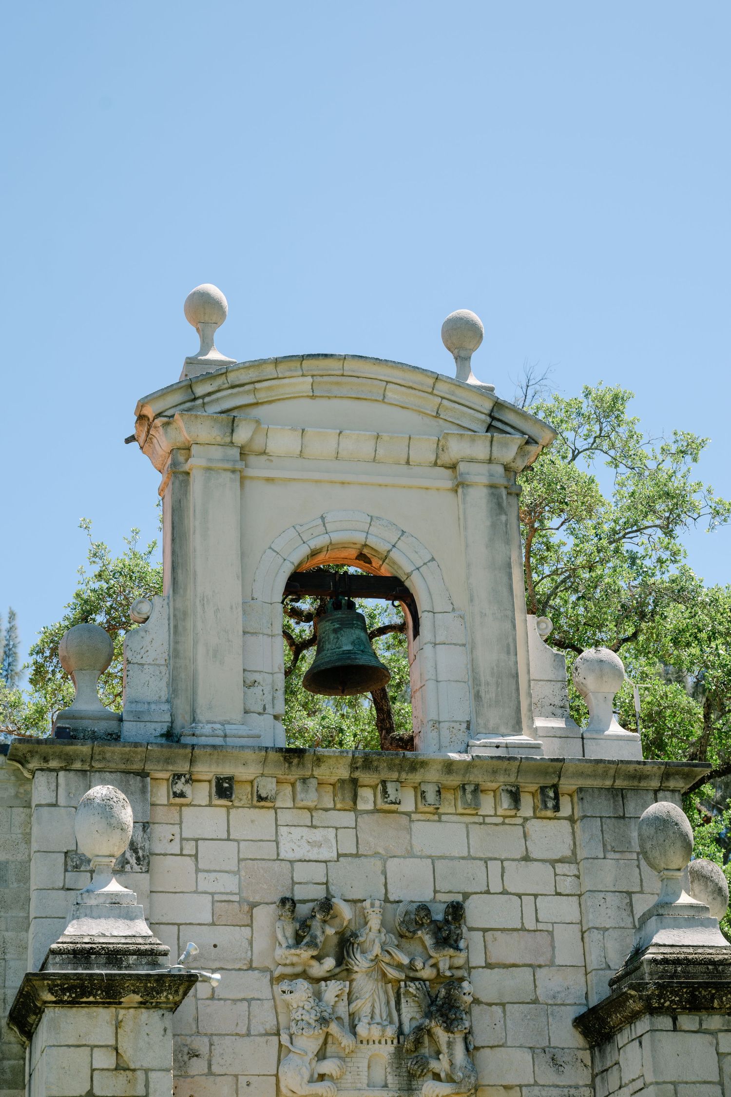A historic stone bell tower with a bronze bell and decorative carvings against a clear blue sky.
