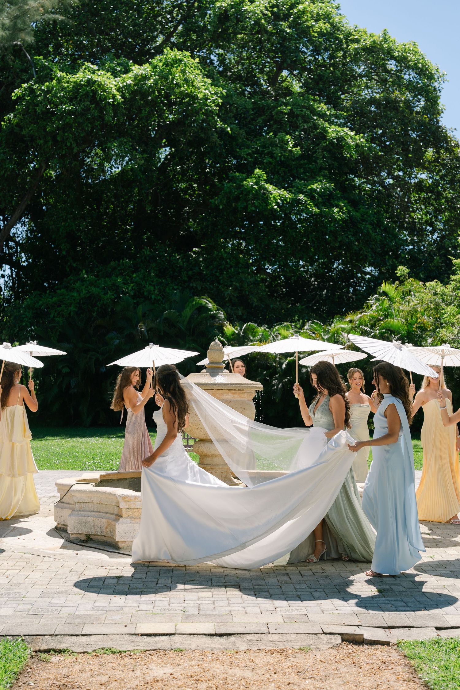 Bride in white gown surrounded by bridesmaids in pastel dresses holding parasols in a lush garden.