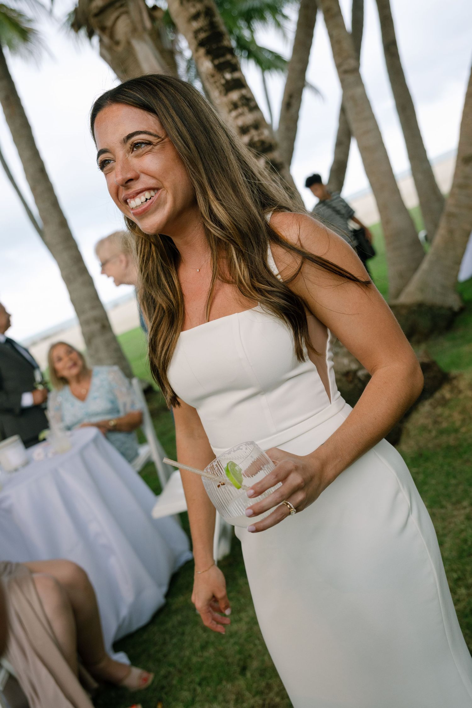 Smiling bride in a white spaghetti strap gown holds a cocktail drink at an outdoor tropical wedding reception.